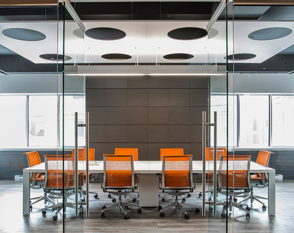 Modern conference room with a glass wall, a long white table, and eight orange office chairs. The room has large windows, gray walls, wood flooring, and a ceiling with round black acoustic panels.