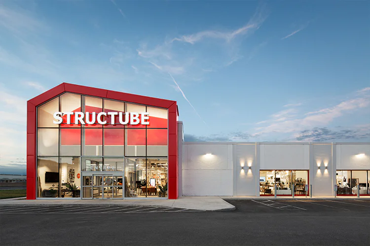 A modern Structube store with a red-framed glass entrance is illuminated at dusk. The building’s large sign and well-lit interior are visible, with empty parking spaces in front and a clear sky overhead.