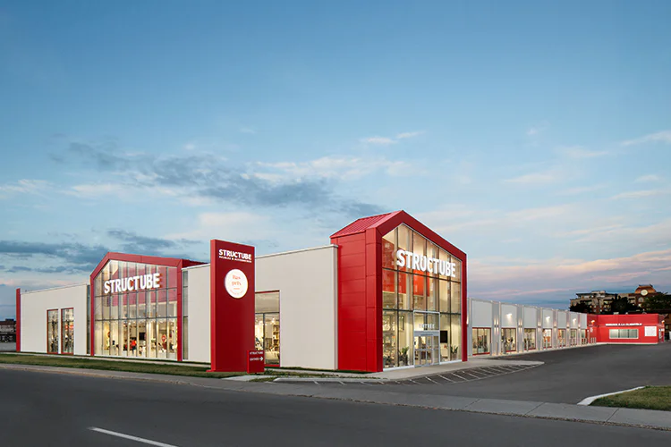 Modern furniture store with white walls and bold red accents, large display windows, and “Structube” signage, located on a corner lot under a blue sky at dusk.
