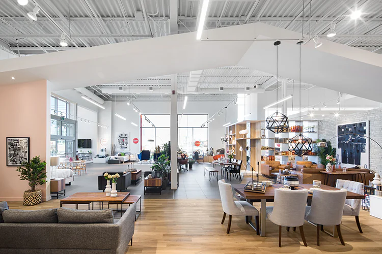 Modern furniture showroom with high ceilings, large windows, and bright lighting. Various sofas, tables, chairs, and decorative items are arranged across wooden and tiled floors. Contemporary decor and plants enhance the open, inviting space.