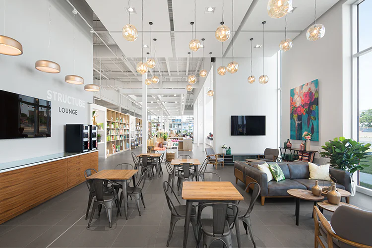 A modern, bright lounge with wooden tables and chairs, a gray sofa with colorful pillows, hanging glass lights, bookshelves, wall art, and large windows letting in natural light.