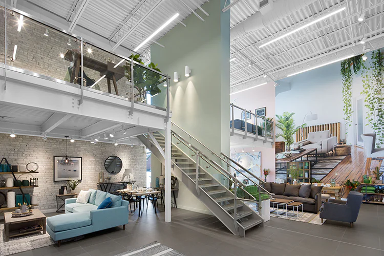 A modern furniture showroom with high ceilings, a mezzanine, glass railings, and stylish sofas, tables, and home decor. Large wall art and plants add a cozy touch. A central staircase connects the two levels.
