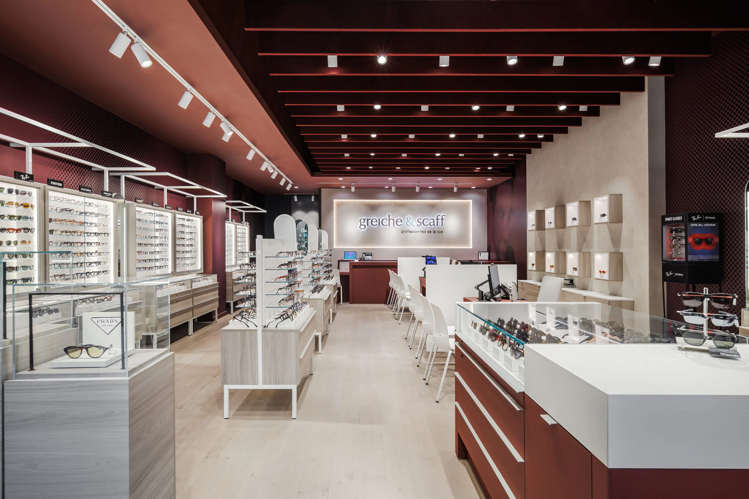 Modern eyewear store interior with display cases of eyeglasses and sunglasses, white chairs at a counter, and illuminated "greiche & scaff" sign on the back wall. The space has red, beige, and white decor with bright lighting.
