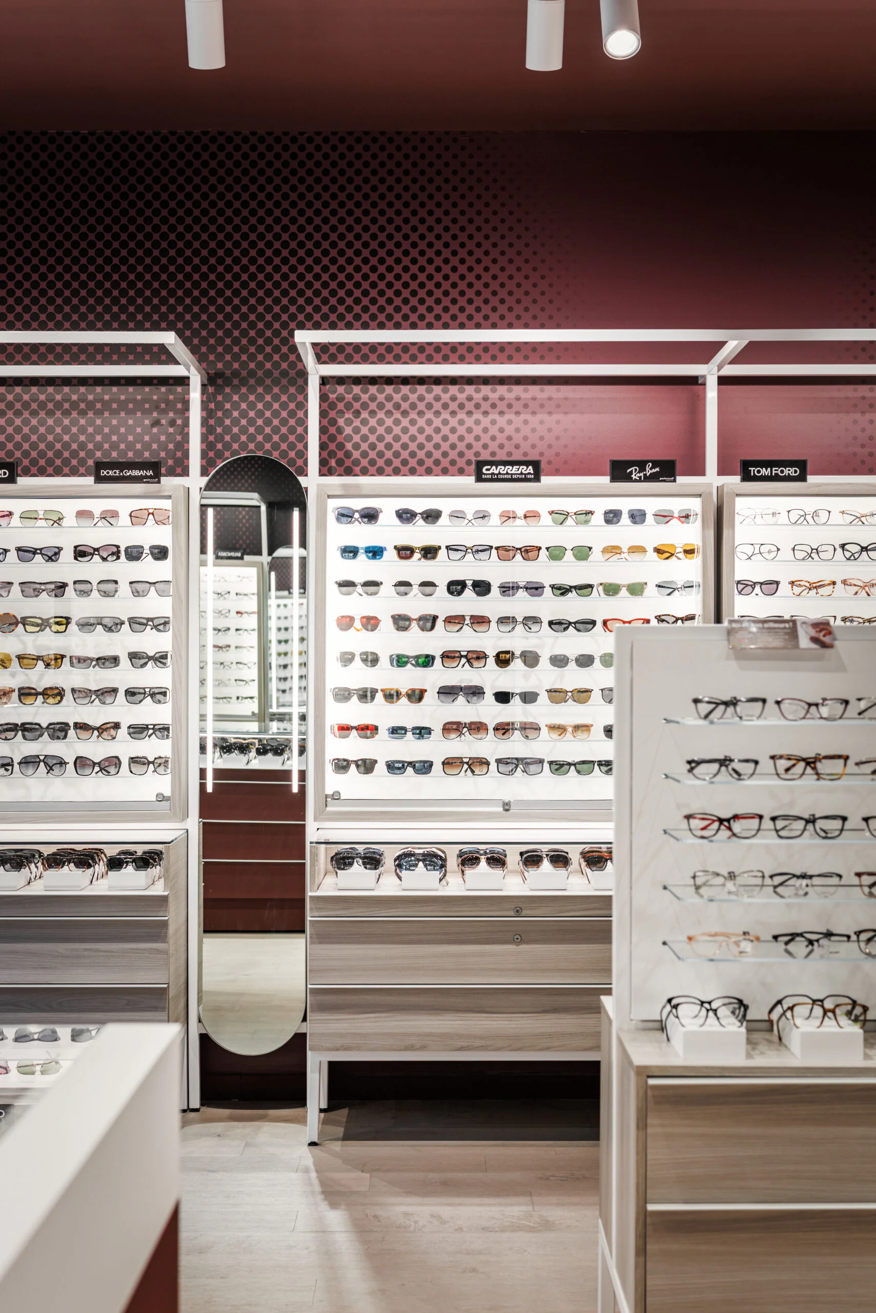 A display wall in an eyewear store features multiple rows of eyeglasses and sunglasses. Signs above showcase brands like Ray-Ban and Tom Ford. The decor includes wood drawers, a mirror, and a maroon accent wall.