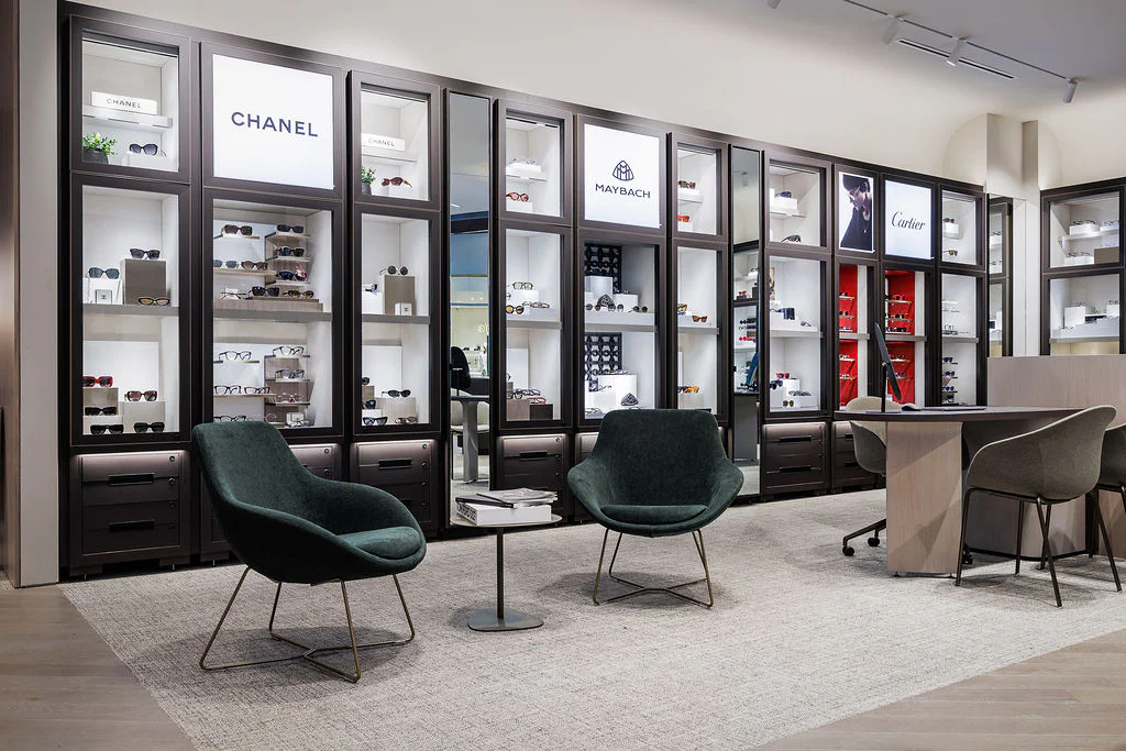 A modern eyewear store interior with display shelves featuring luxury brands like Chanel, Maybach, and Cartier. Two dark green chairs and a small table sit on a gray rug in front of the displays.