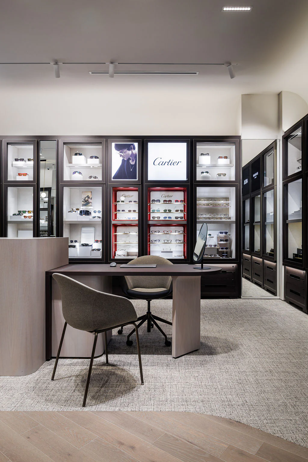 A modern eyewear boutique features a sleek desk with two chairs, display shelves of designer glasses, soft lighting, and a prominent Cartier sign on the back wall.