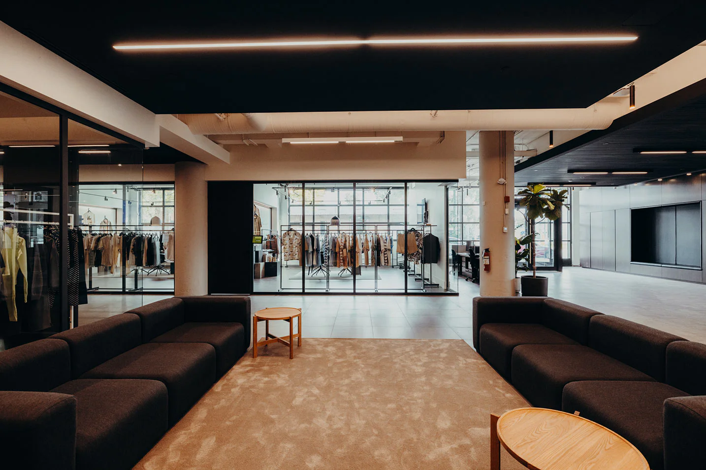 Modern office lounge with dark couches, a beige rug, and small wooden tables. Glass walls reveal a clothing display and racks in an adjacent room. Large windows let in natural light.