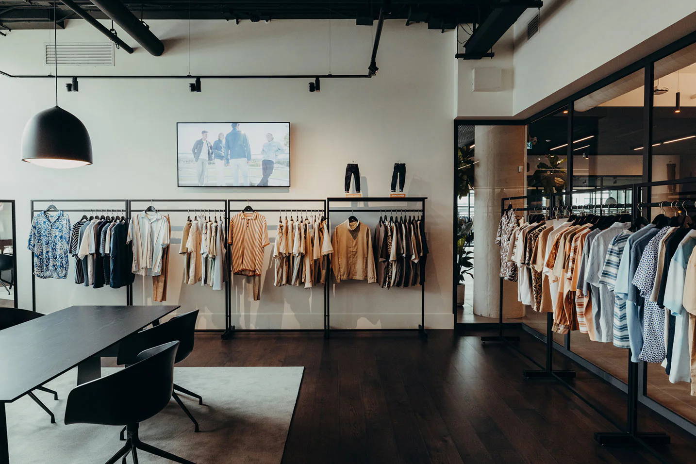 Modern clothing showroom with racks of neatly arranged men's shirts and jackets in neutral tones, a large black table with chairs, and a digital screen displaying a fashion image on a white wall.