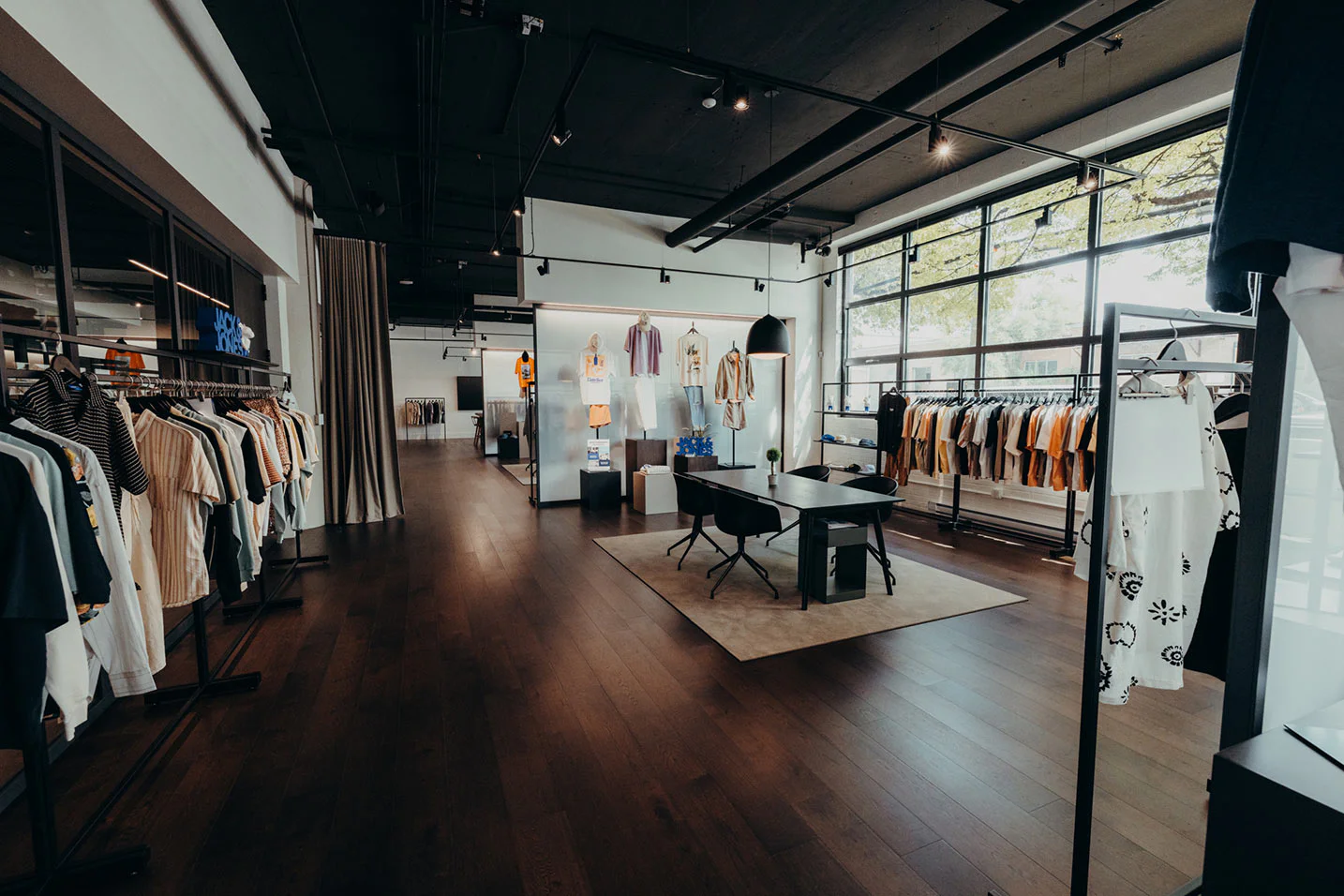 A modern clothing boutique with wooden floors, large windows, and racks of neatly arranged clothes. A desk with chairs sits in the center, and natural light fills the spacious, stylish interior.