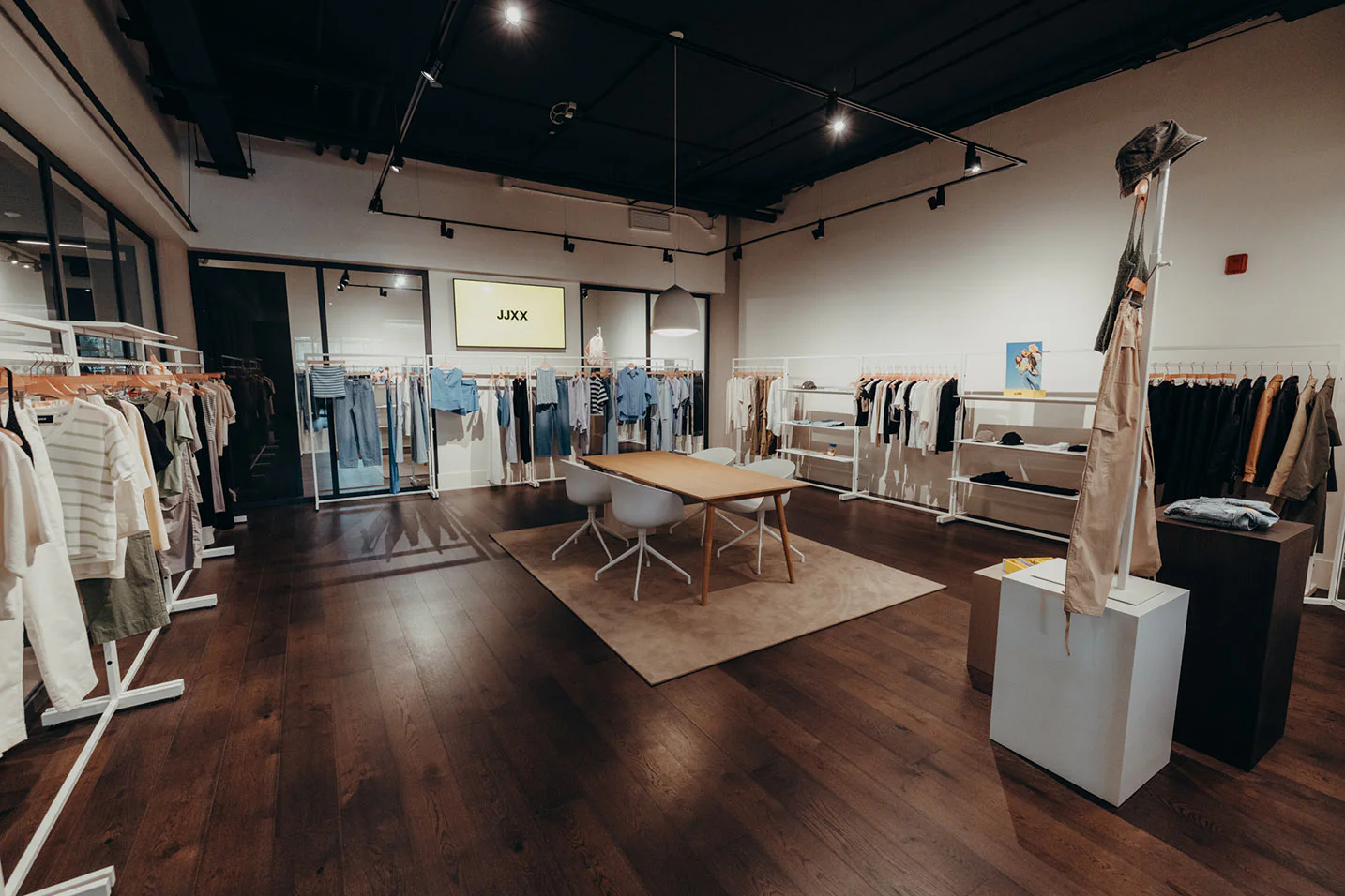 A modern clothing store interior with wooden floors, minimalist racks displaying neutral and pastel clothes, a central table with chairs, and a wall-mounted screen displaying the brand name "JIXY.