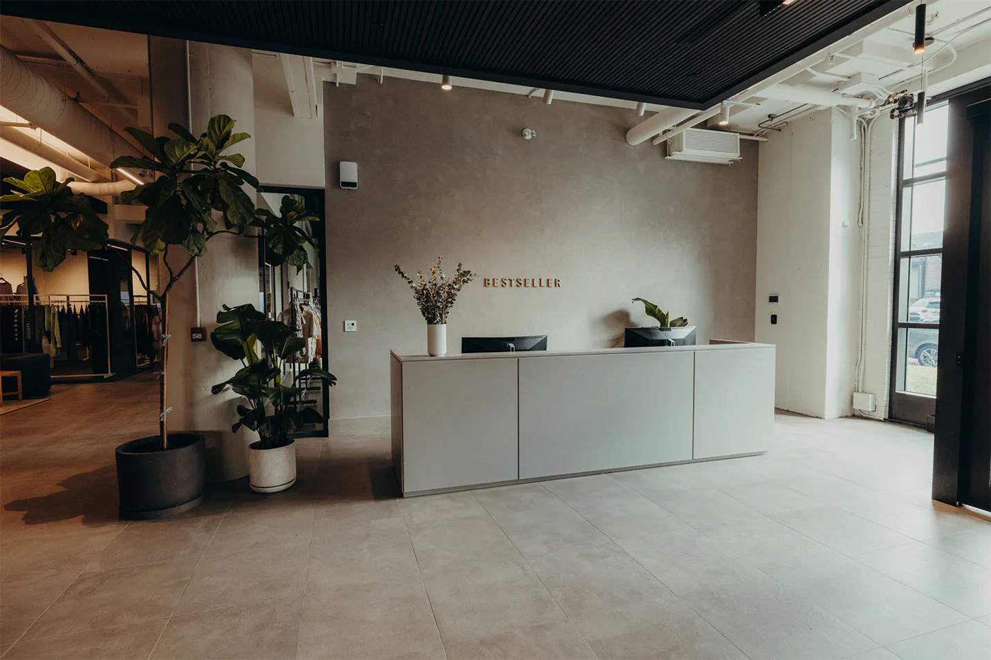 A modern, minimalist office reception area with a gray desk, two computer monitors, potted plants, and a wall sign reading "Bestseller." Large windows let in natural light.