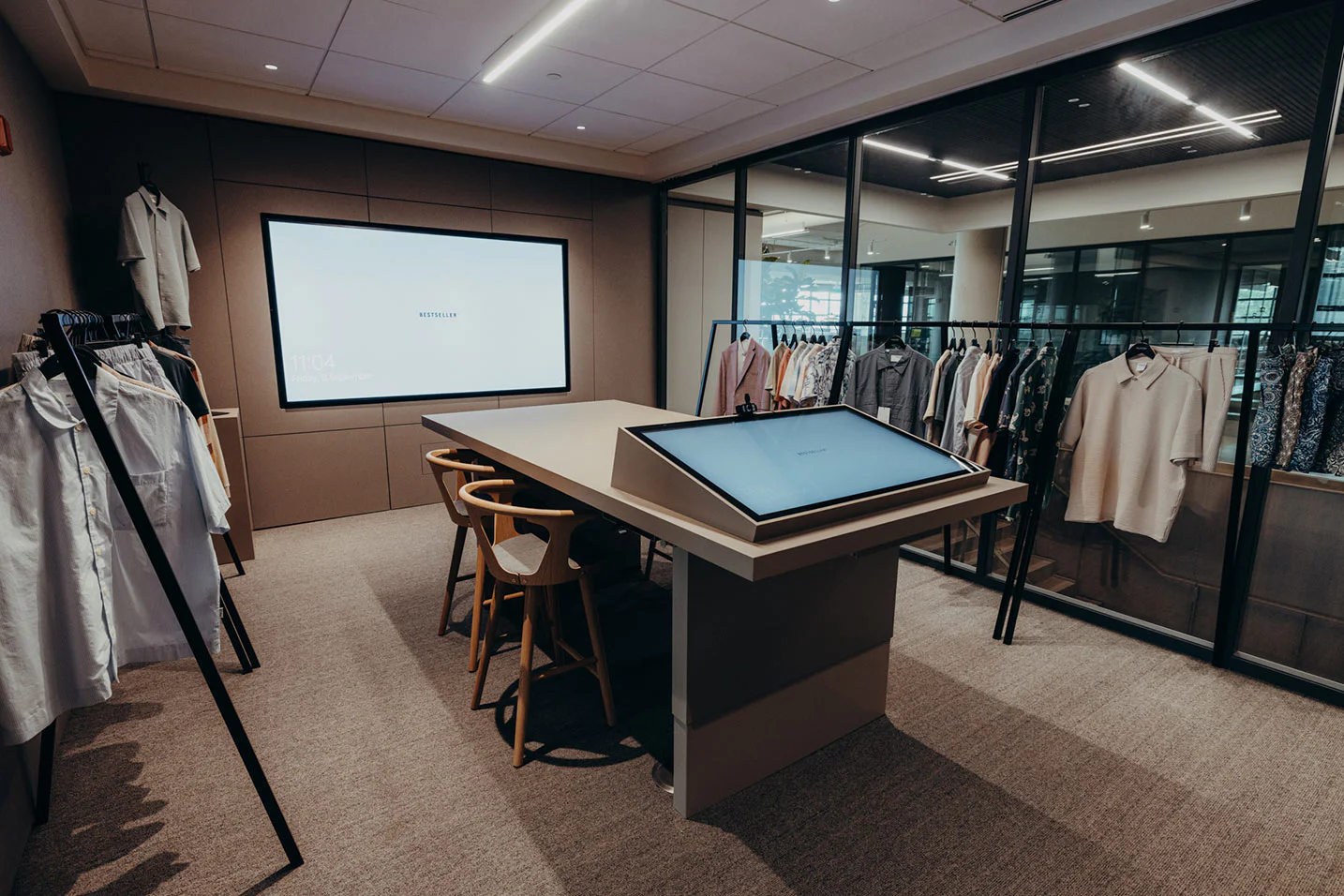 A modern showroom with racks of clothing, a large touchscreen table, wooden chairs, and a digital display on the wall, set in a sleek, well-lit office space.
