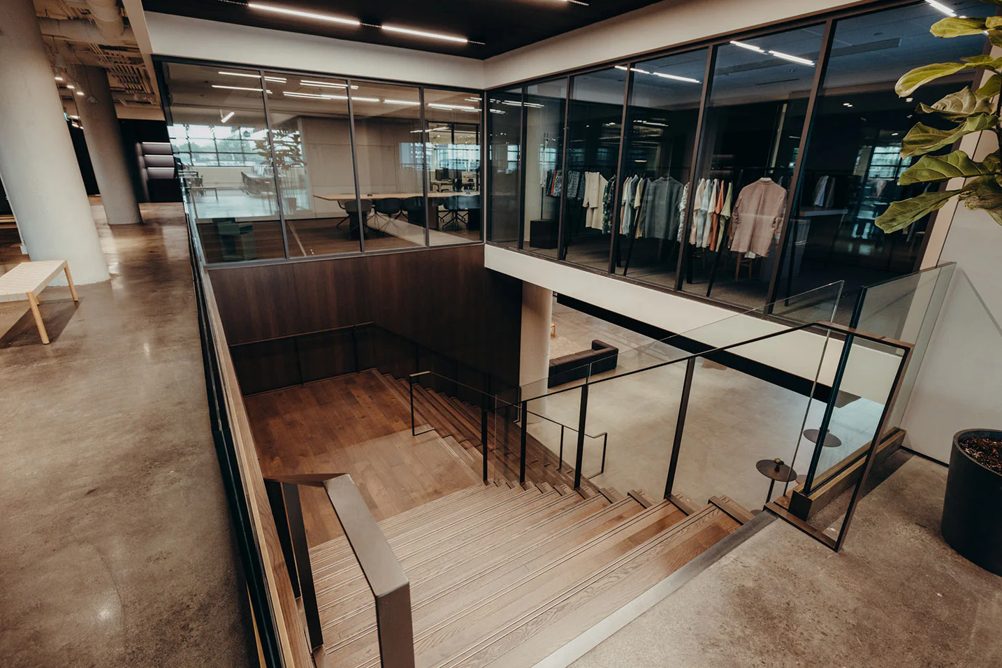 A modern office interior with glass walls, a wooden staircase, polished concrete floors, and a room displaying hanging clothes visible through the windows on the upper floor.