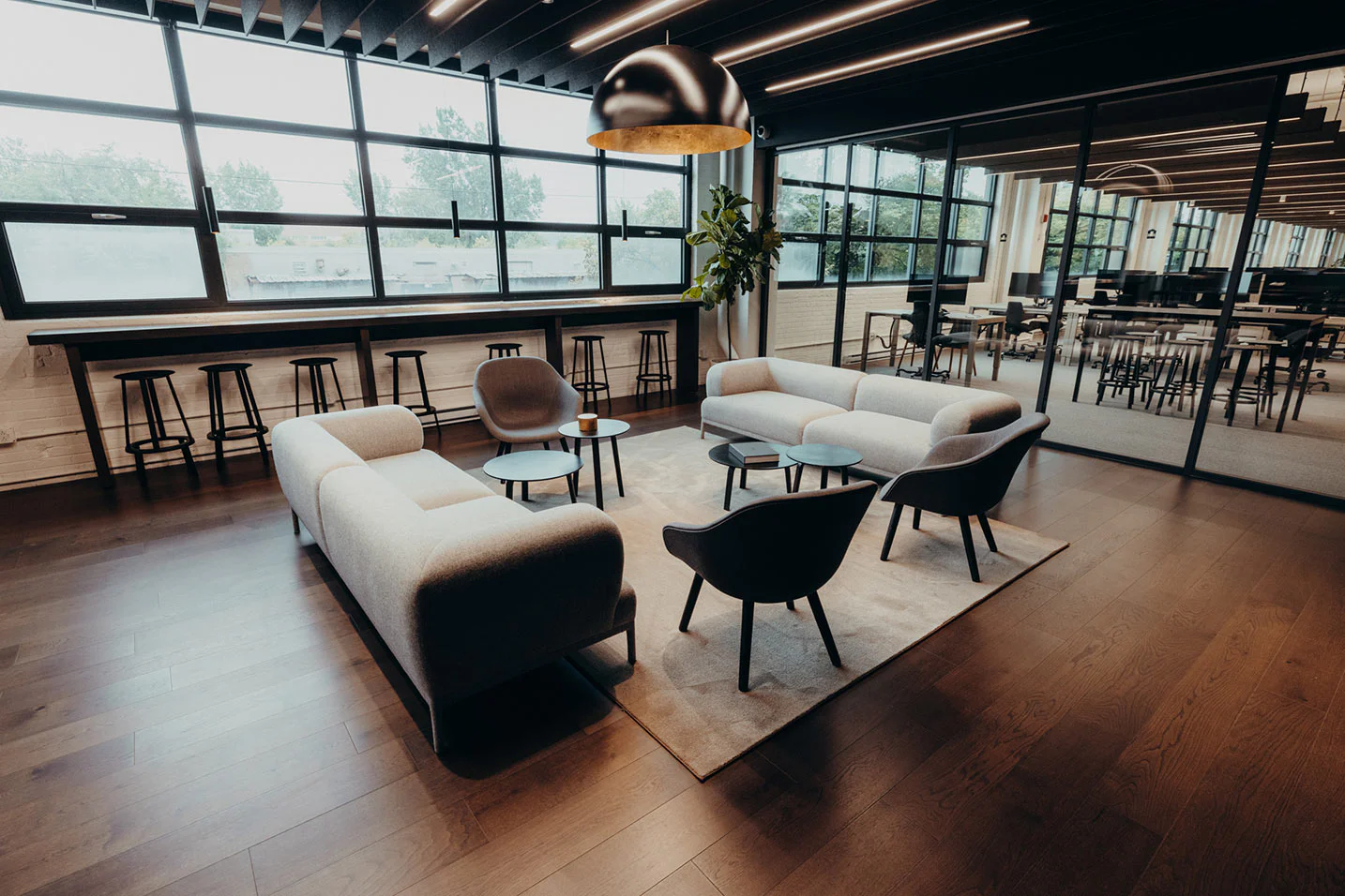 Modern office lounge with two gray sofas, four black armchairs, small round tables, a large rug, and a gold hanging lamp. Large windows and glass walls let in natural light; barstools line a counter by the window.