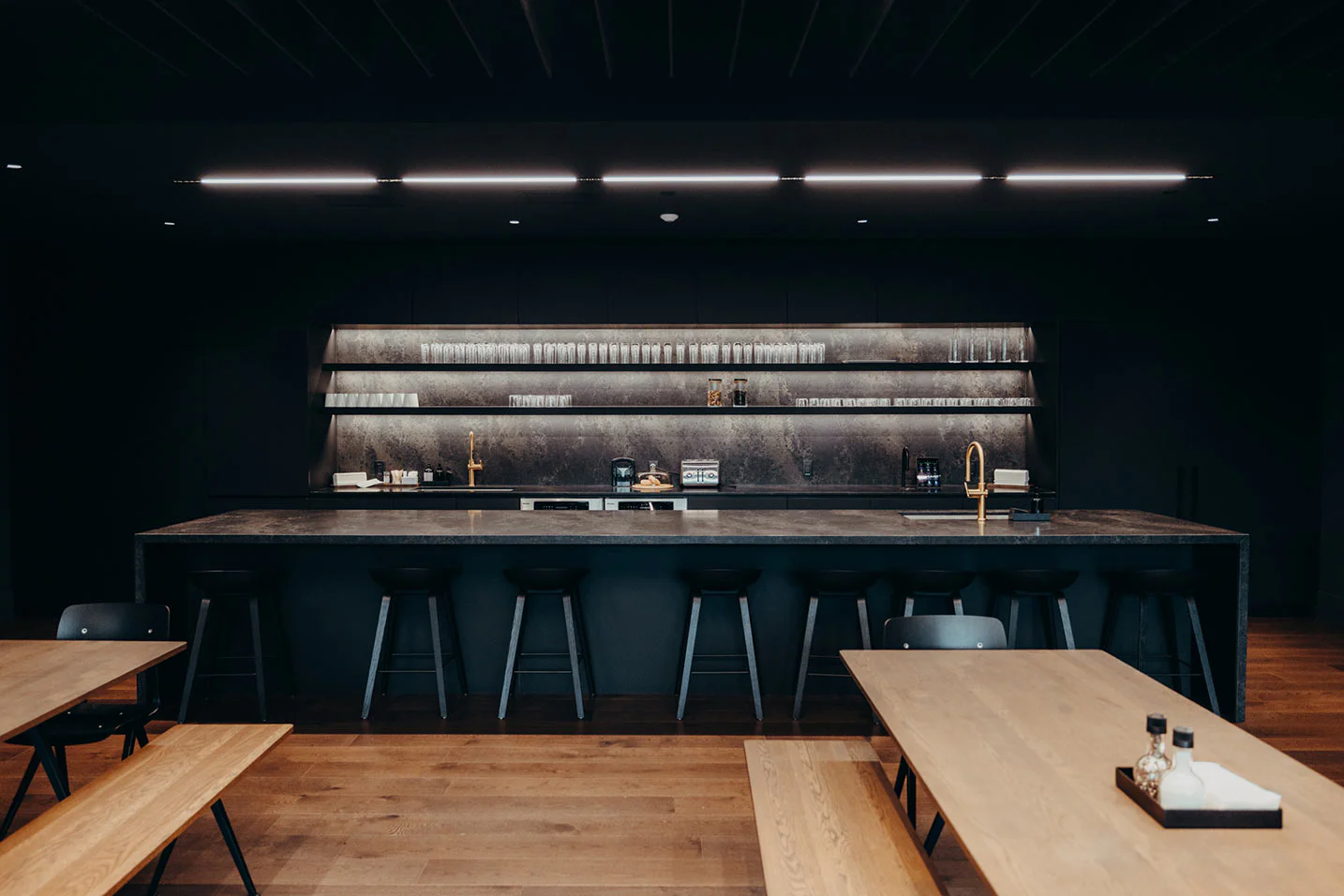 A modern, minimalist bar with a long concrete counter, black stools, open shelves with glassware, wooden tables and benches, and dim, warm lighting. The overall color palette is dark and neutral.