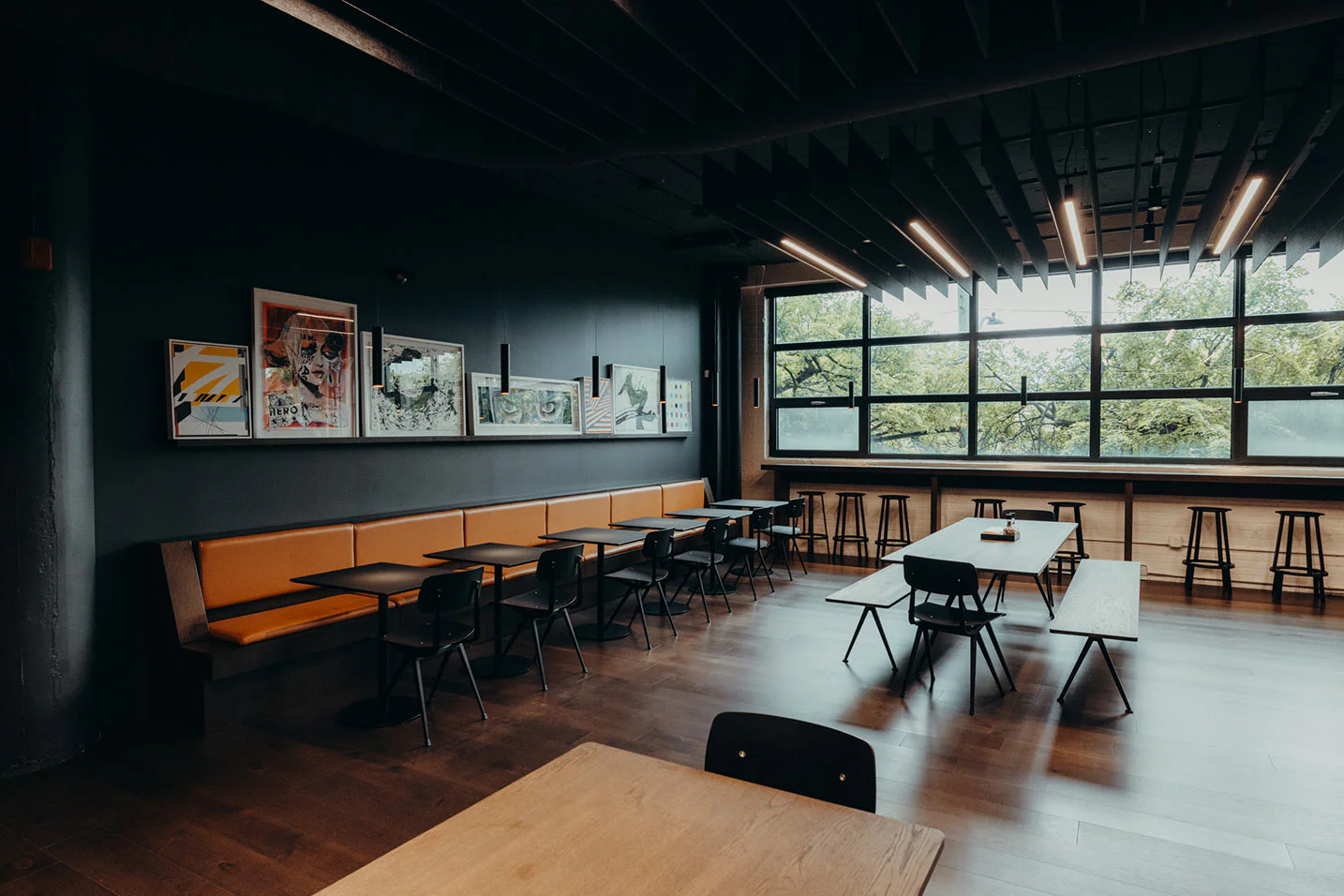 A modern, empty café with wooden floors, black chairs and tables, framed artwork on a dark wall, a long cushioned bench, and large windows letting in natural light with trees visible outside.