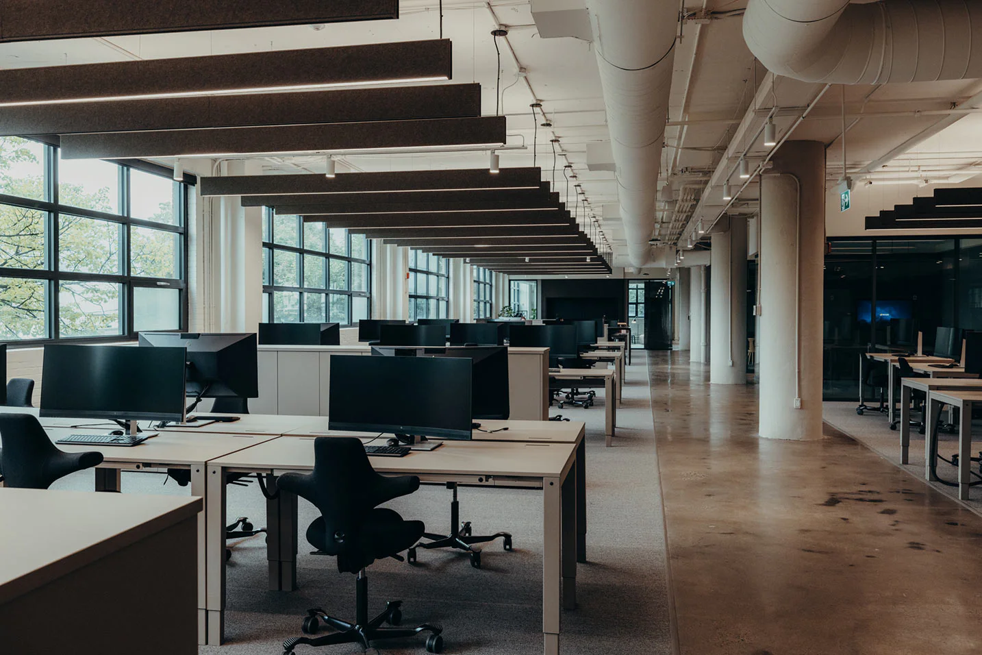 A modern, empty open-plan office with rows of desks, office chairs, and computer monitors. Large windows line one side, letting in natural light. The space features exposed ceilings and industrial-style lighting.