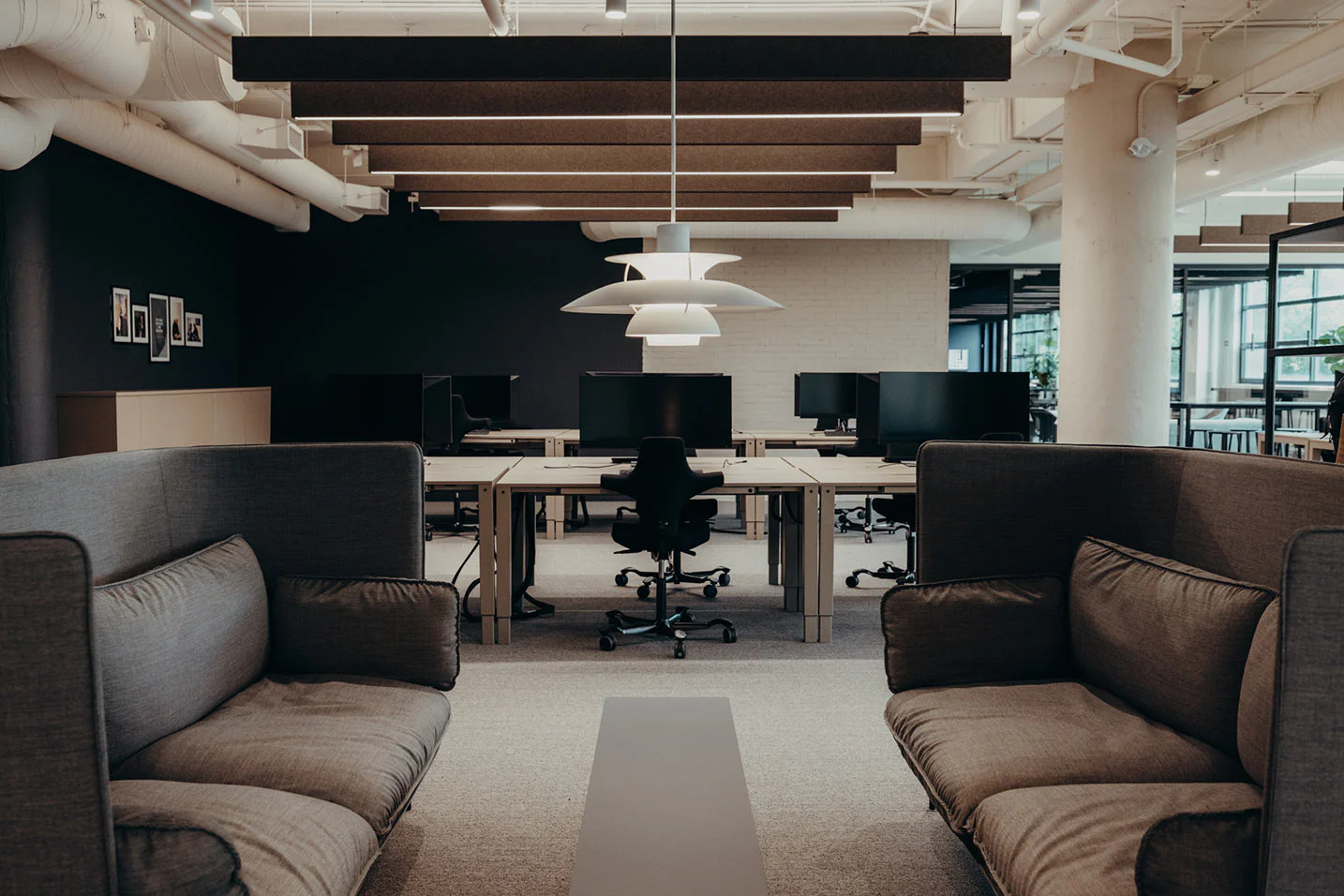 A modern, open-plan office with gray sofas facing each other, desks and office chairs in the background, multiple monitors, and a white hanging lamp above a coffee table. The space is well-lit with natural and artificial light.