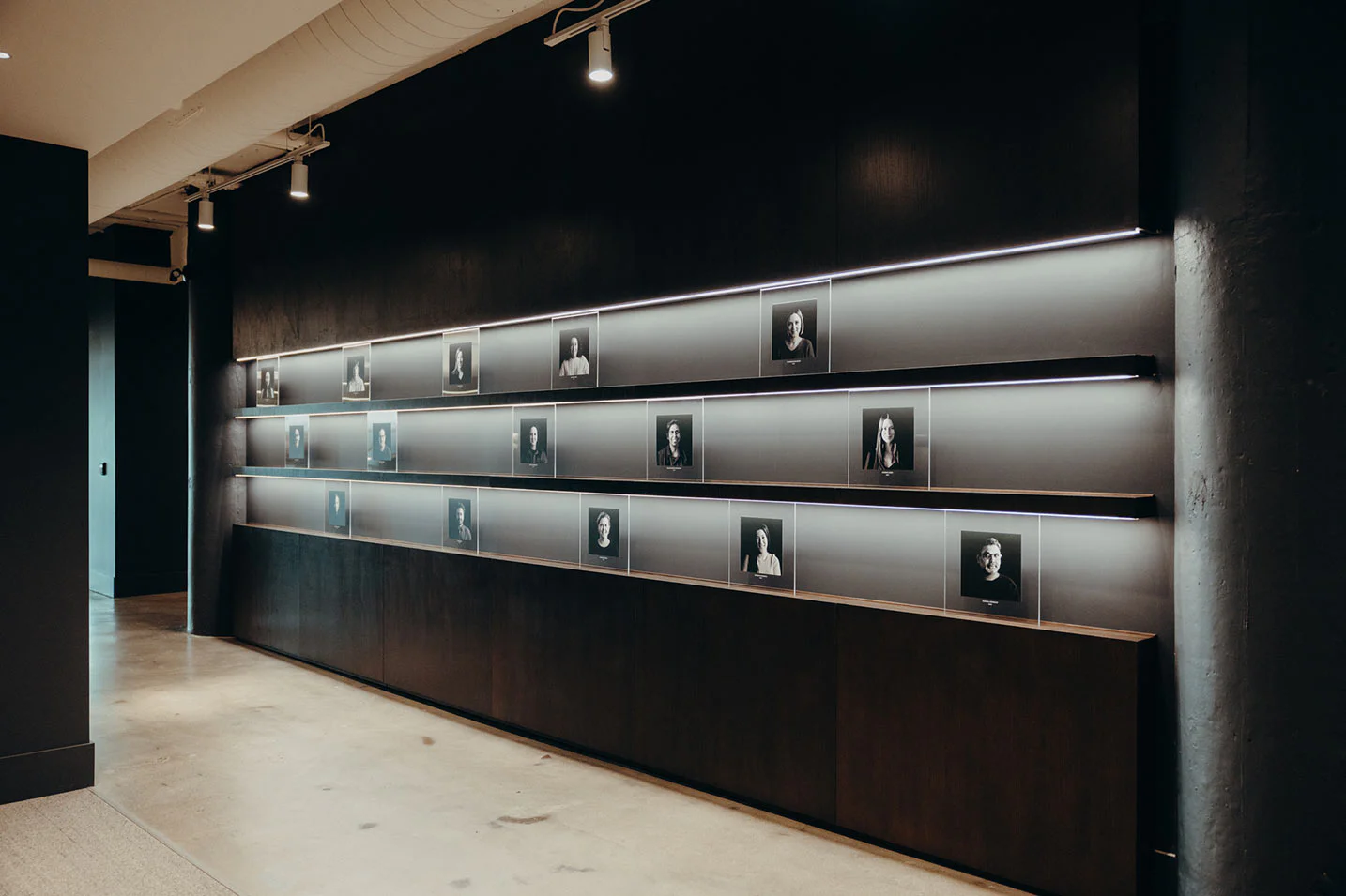 A modern hallway features a dark wall with horizontal shelves displaying rows of black-and-white portrait photographs, each illuminated by sleek overhead lighting. The floor is polished concrete.