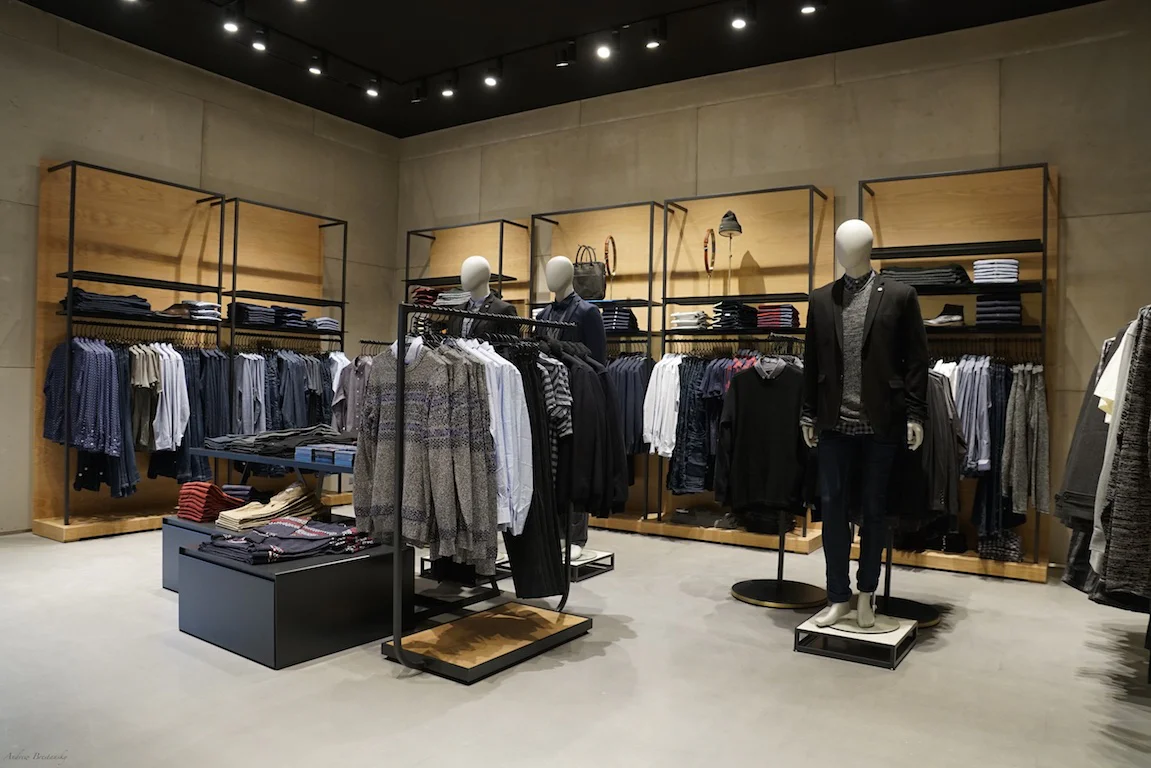 A modern clothing store with wooden shelves and black racks displaying neatly arranged shirts, pants, and jackets. Two mannequins are dressed in casual outfits, and folded clothes are stacked on tables. The store has a minimalist design.