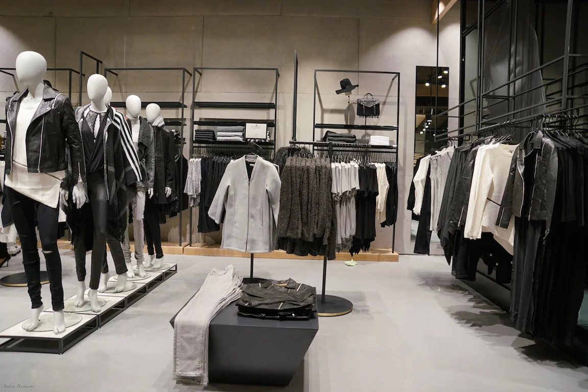 A modern clothing store interior with mannequins dressed in black, white, and gray outfits. Racks display neatly arranged shirts, jackets, and pants in neutral tones. The setting is minimalist and well-lit.