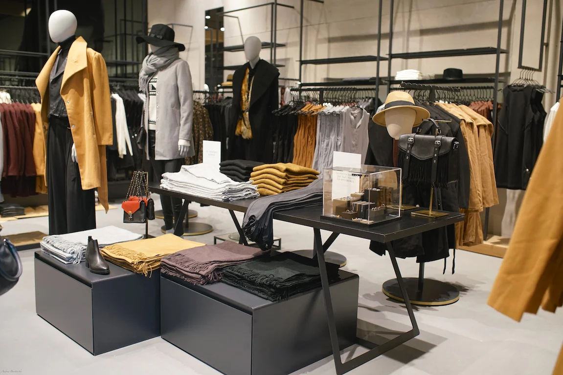 A clothing store display featuring neatly folded sweaters, scarves, and accessories on tables, with racks of hanging coats, jackets, dresses, and mannequins dressed in stylish outfits in the background.