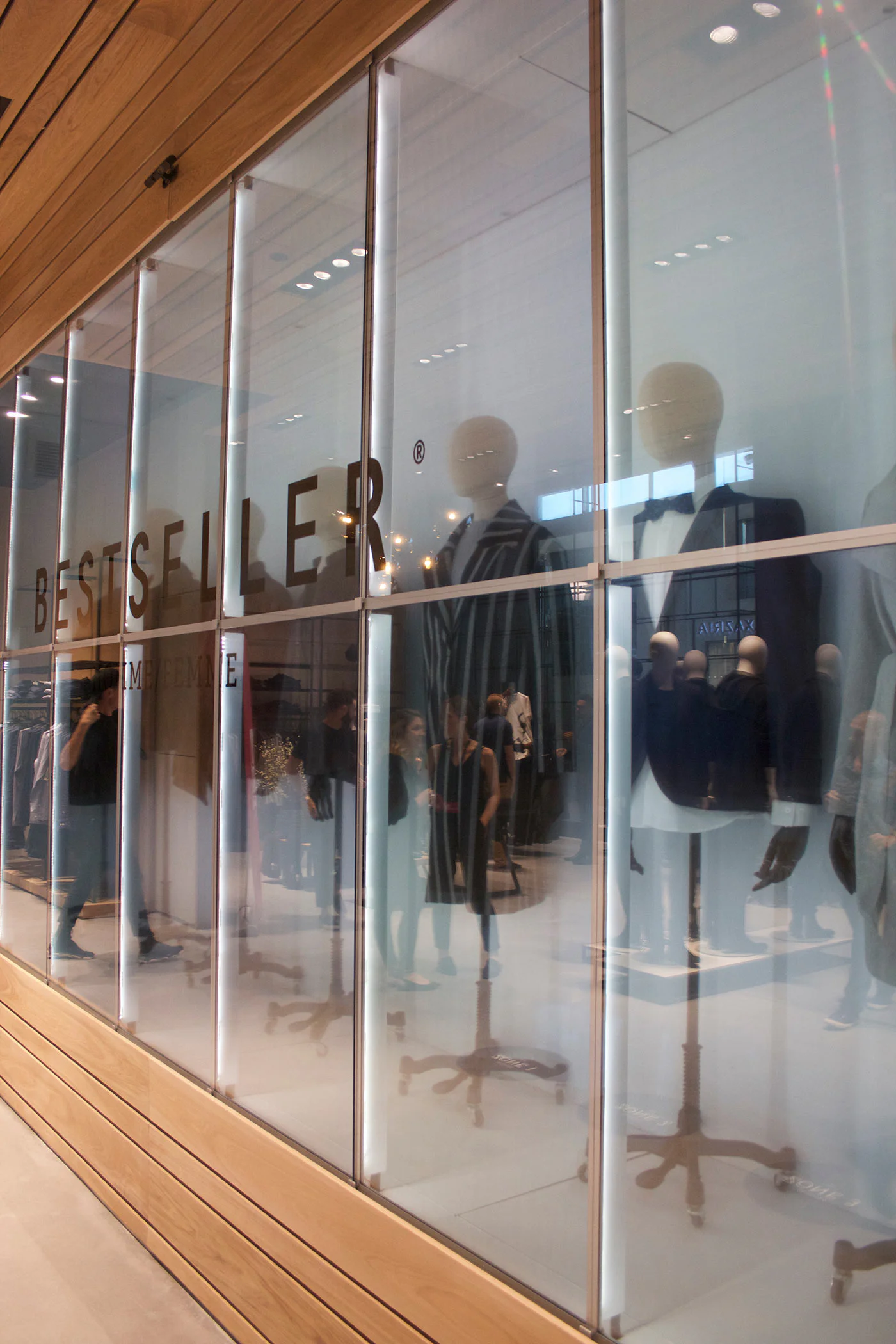 Storefront window display with the word "BESTSELLER" showing several mannequins dressed in formal clothing. The store interior and shoppers are visible through the glass reflection.