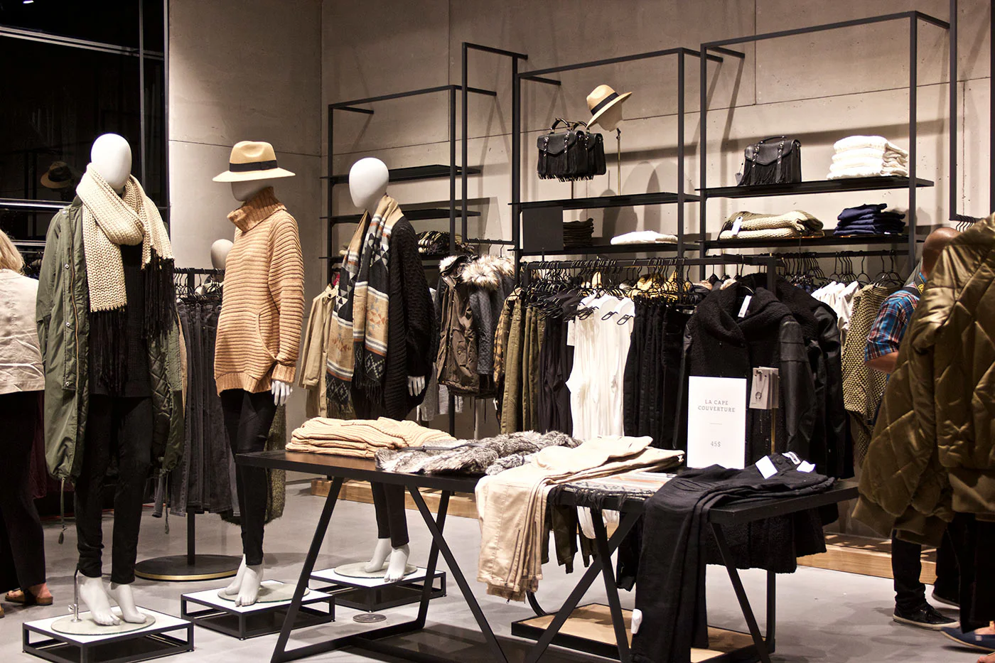 A clothing store displays mannequins dressed in cozy winter outfits, with scarves, hats, and sweaters. Racks and shelves hold neatly folded clothes and accessories, creating a modern and organized shopping space.