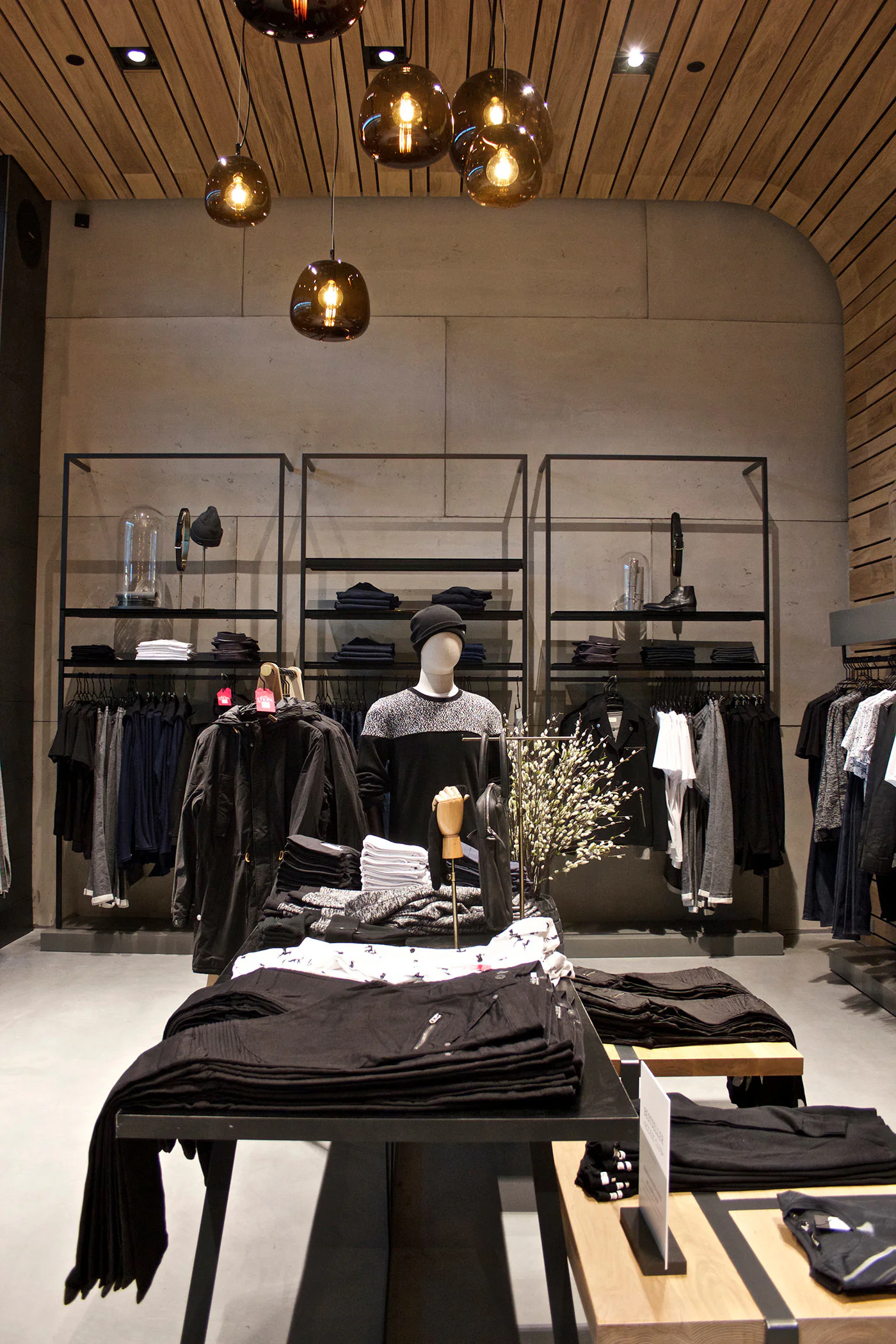 Modern clothing store interior with minimalist decor, featuring folded clothes on wooden tables, a mannequin dressed in black and gray, and shelves with neatly arranged clothing against a concrete and wood-paneled wall.