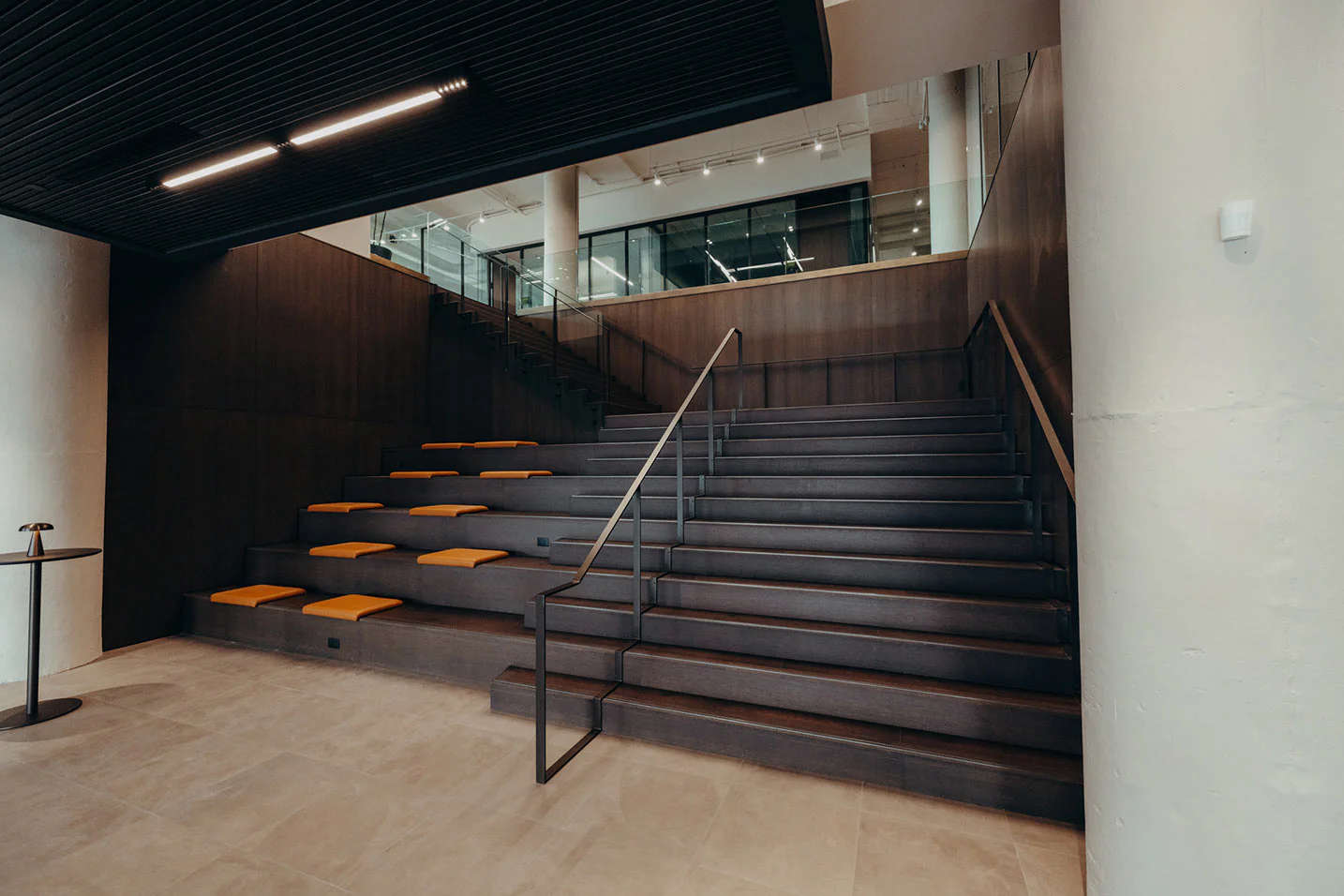 Modern indoor staircase with dark steps and a metal handrail, alongside tiered seating with orange cushions. Glass railings and an open second floor are visible in the background. The space has a minimalist design.