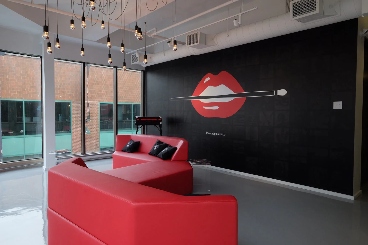 A modern lounge with red sofas, large windows, and a black wall featuring a mural of red lips with a makeup brush. Exposed bulbs hang from the ceiling, and natural light fills the space.