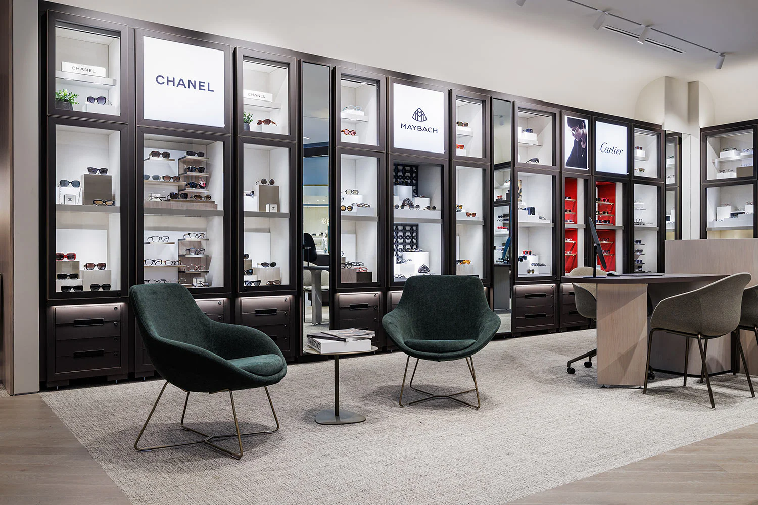 Modern eyewear boutique with glass display cases showcasing designer brands like Chanel, Maybach, and Cartier. Two dark green chairs and a small table sit on a gray rug, creating a stylish seating area.
