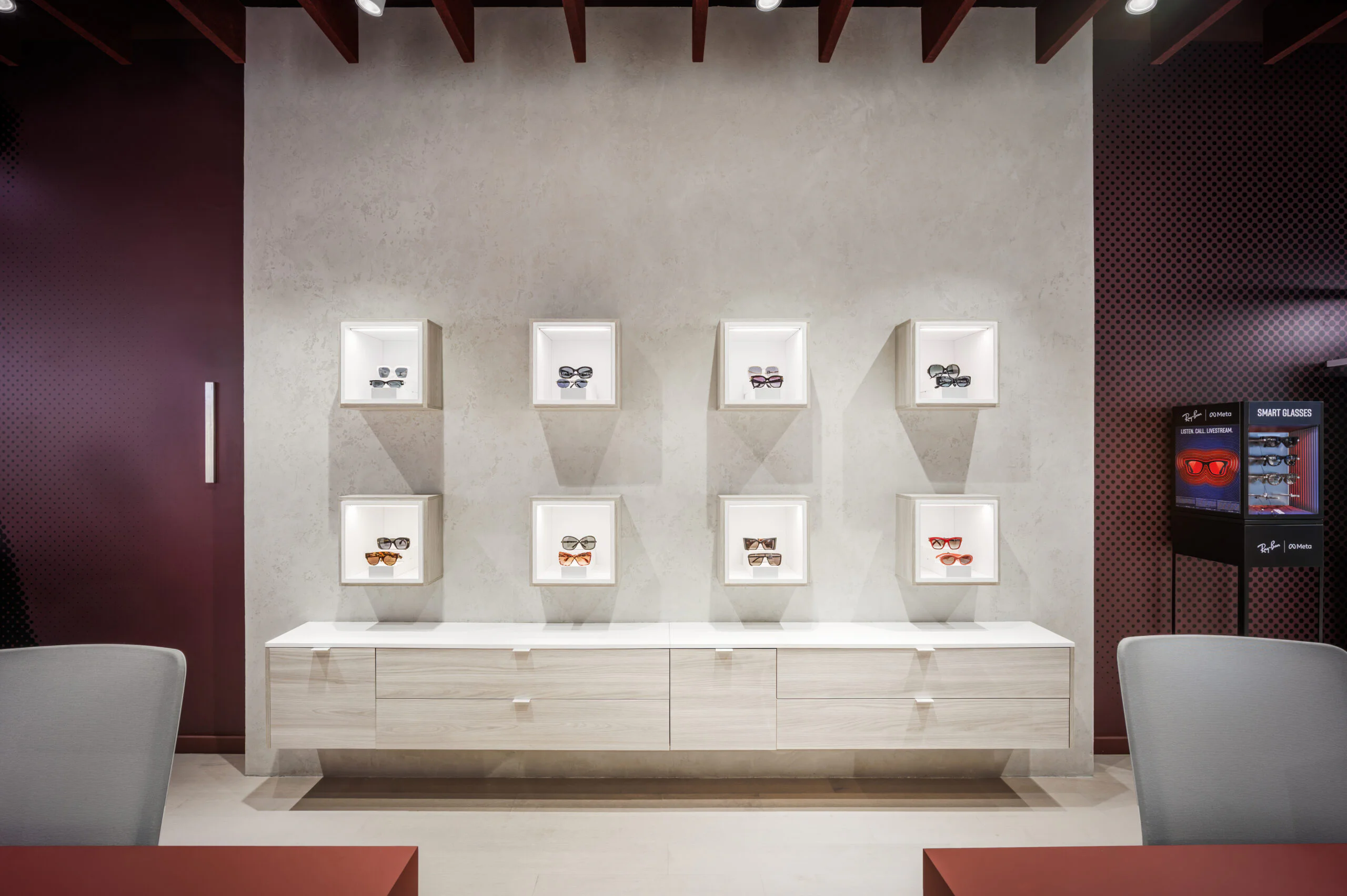 A modern eyewear store display with nine pairs of glasses showcased in illuminated cubed shelves on a light wall, above a white cabinet, with maroon accents and chairs in the foreground.