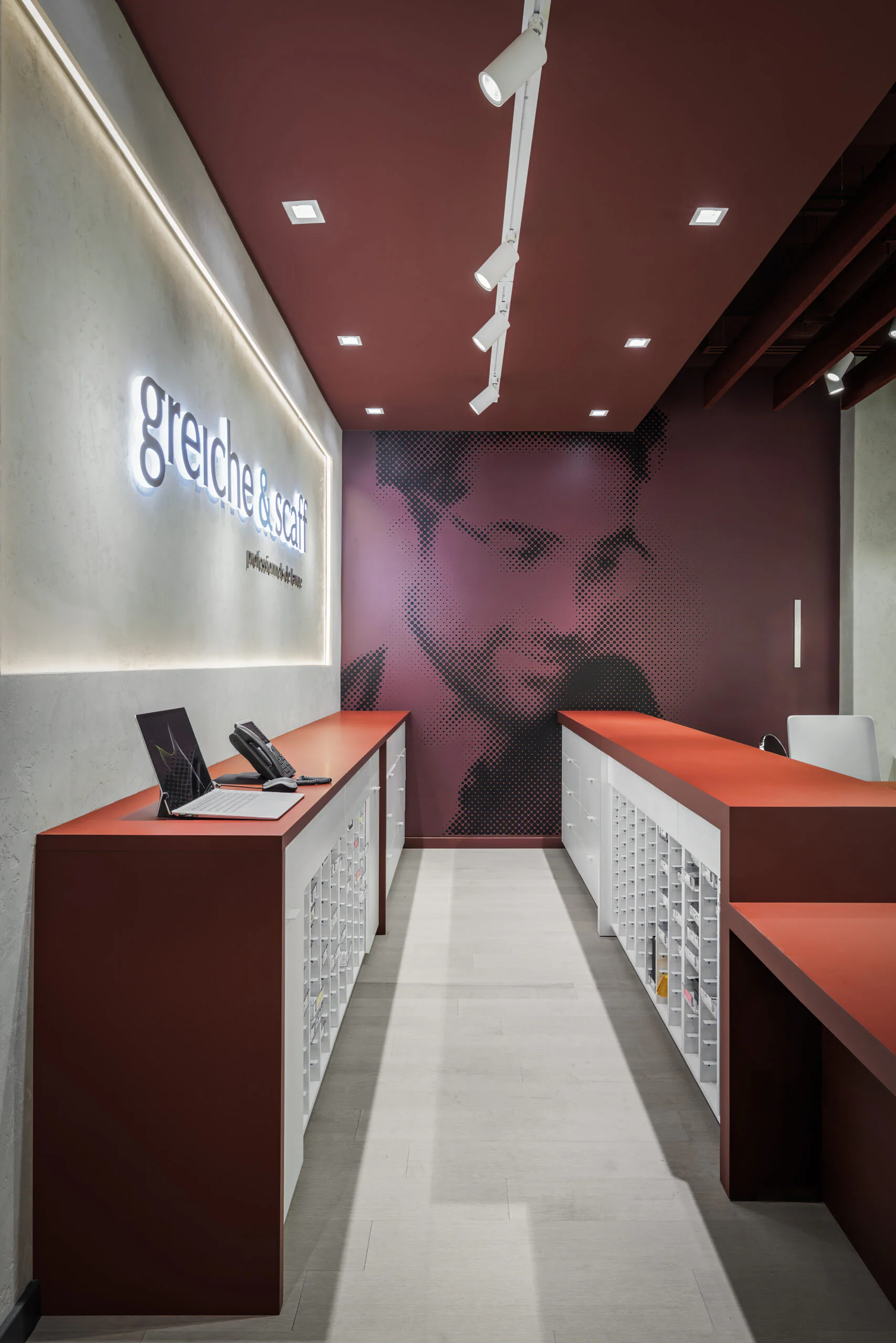 A modern optician’s store interior with red counters, a white wall with “greiche & scaf” signage, and a large photo of a man with glasses on the back wall. Shelves and tablets are arranged on the counters.