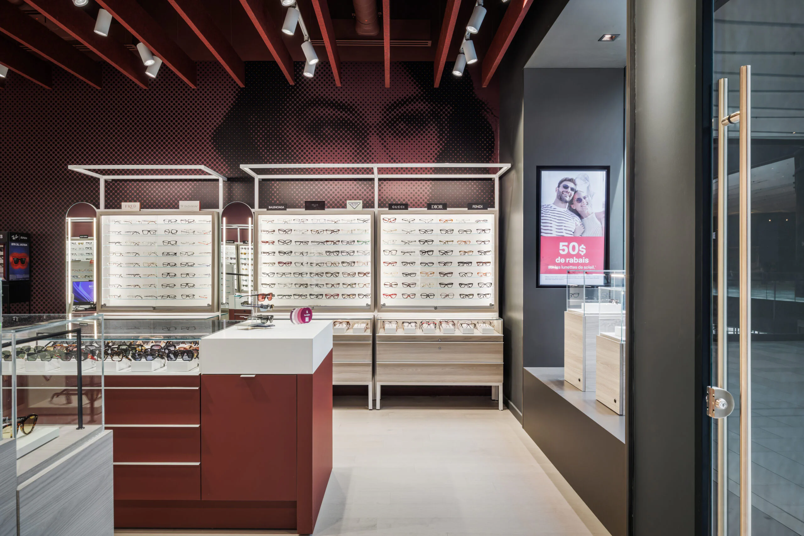 Modern eyewear store interior with red and white decor, display shelves filled with glasses, a central counter, and a digital sign advertising “50% de desconto” near the entrance. Large wall art of a woman's face in the background.