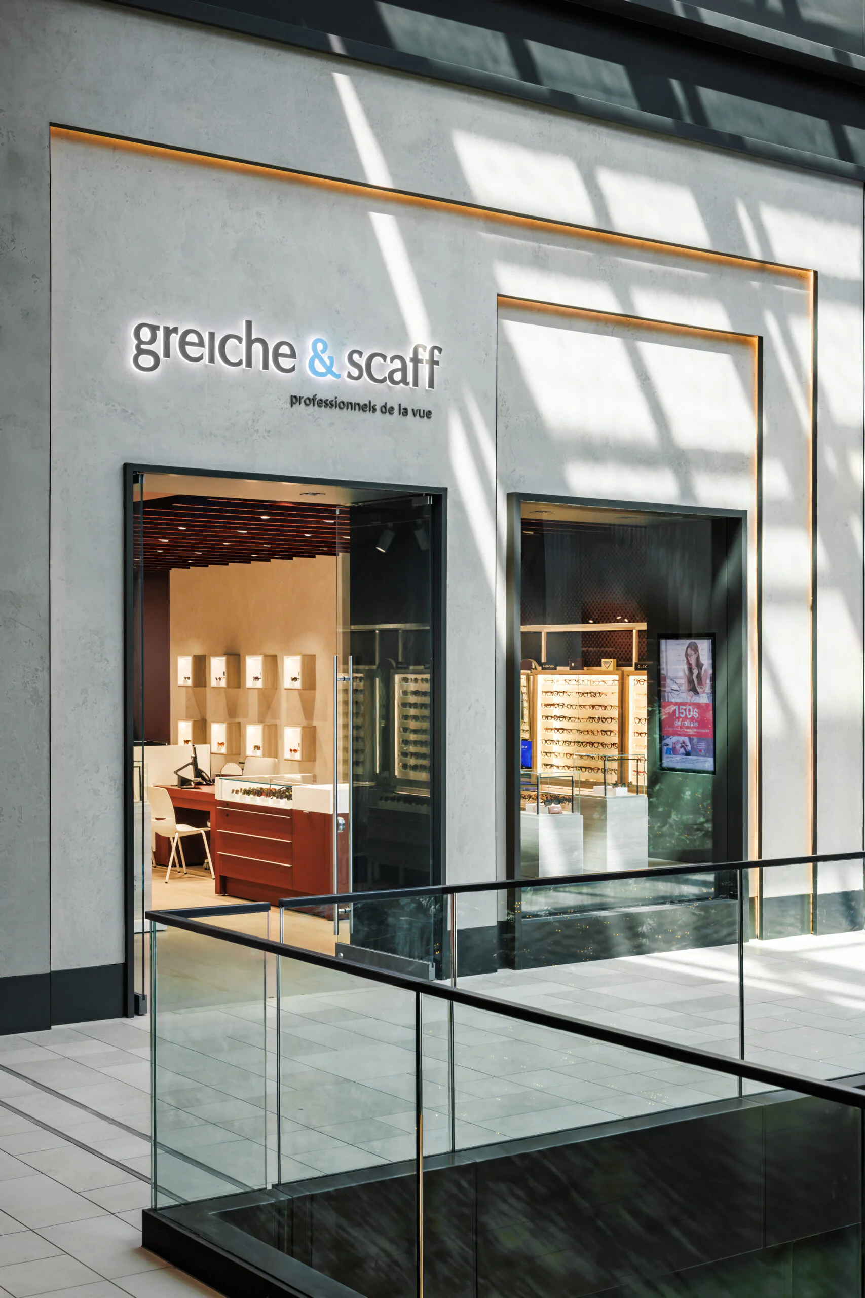 A modern optician store named "greiche & scaff professionnels de la vue" is in a shopping mall, with display shelves and eye care products visible through large windows. Sunlight casts shadows on the floor and walls.