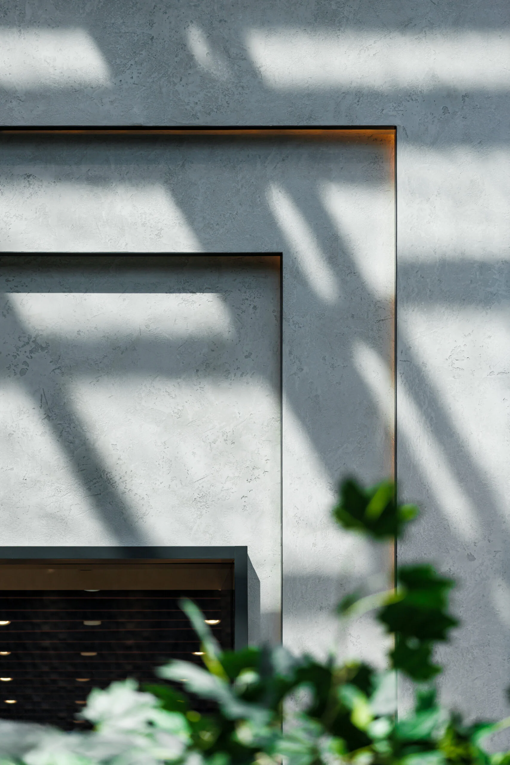 Sunlight casts geometric shadows on a textured gray wall with rectangular recesses. Out-of-focus green leaves appear in the lower foreground, adding a natural element to the modern, minimalist scene.
