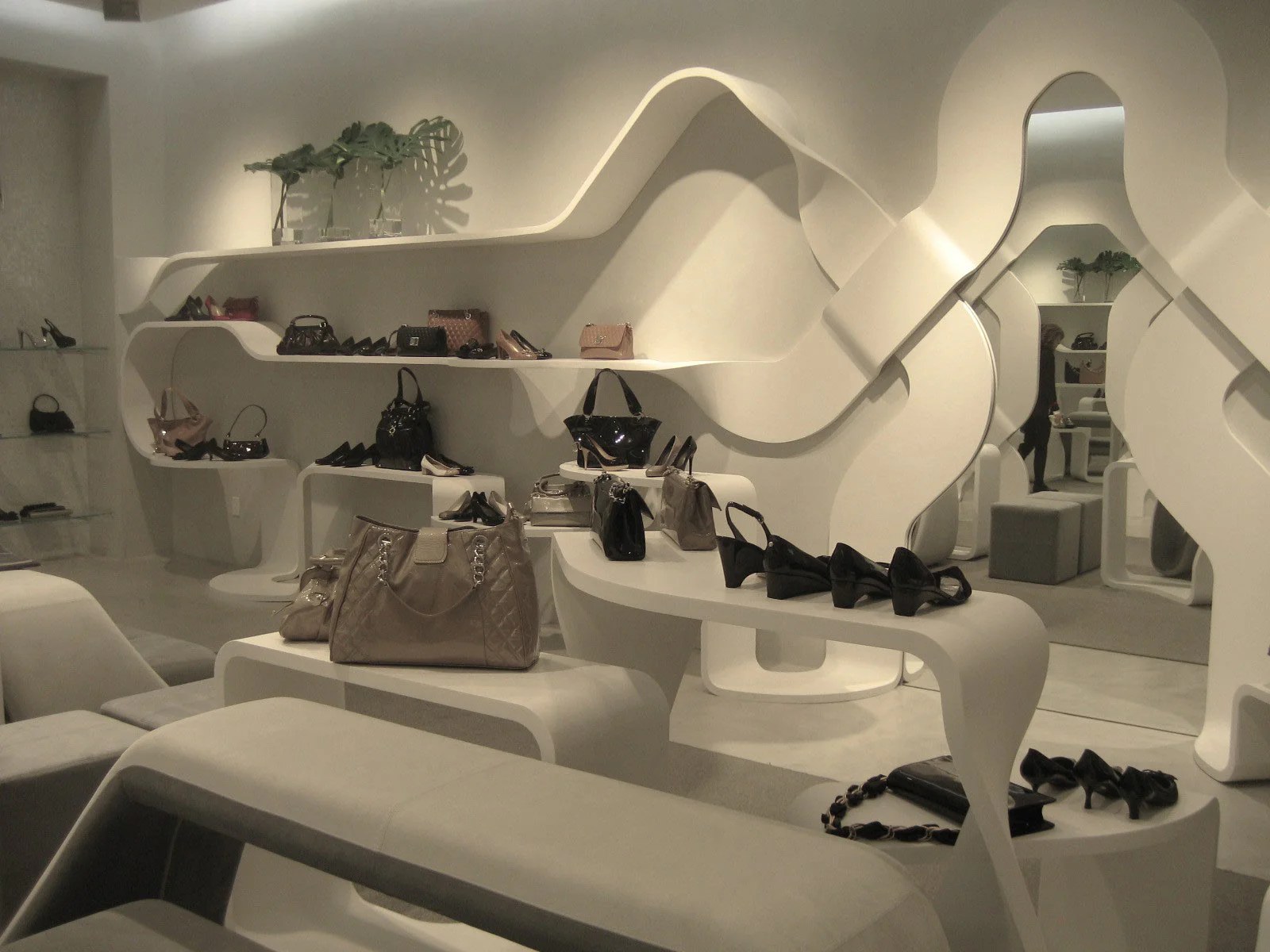 A modern boutique with white, sculptural shelves and tables displaying handbags and shoes. The space has curved, futuristic designs, minimalist decor, and a few green plants on a top shelf.