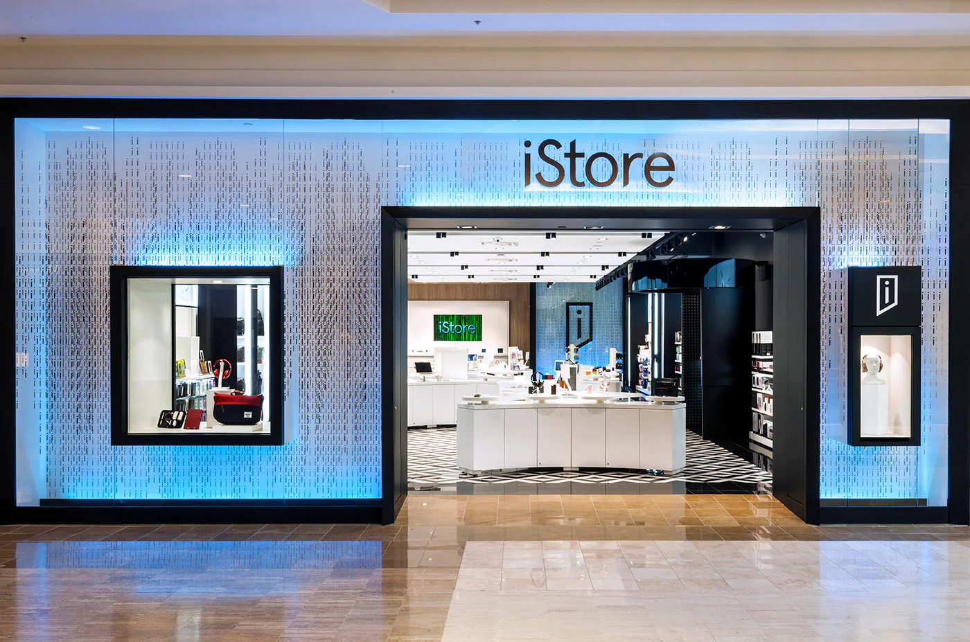 A modern iStore with a blue-lit exterior, showcasing tech products in window displays and a bright, organized interior visible through large glass entrance doors.