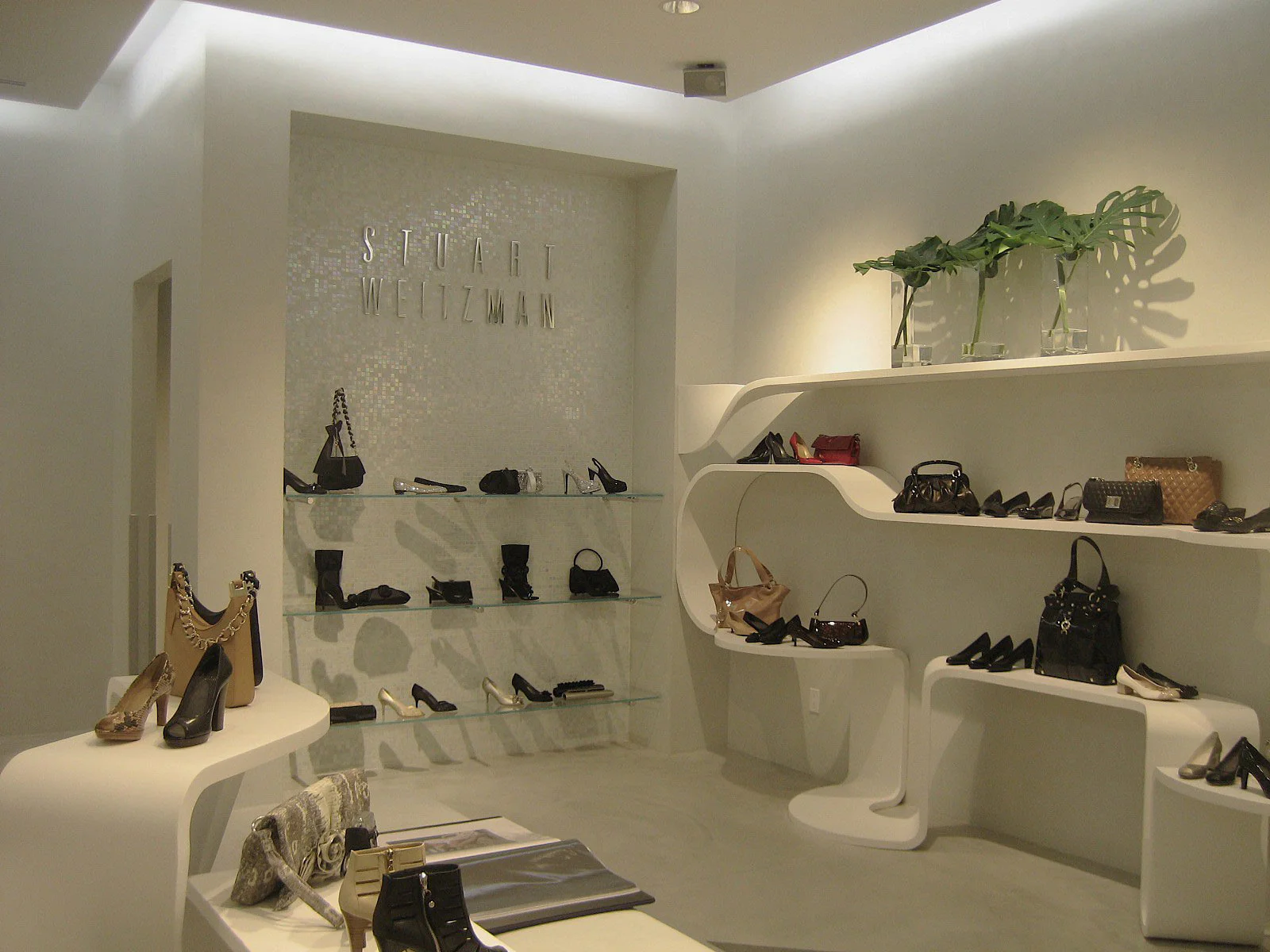 A modern shoe store interior displays various high-heeled shoes and handbags on white curved shelves and glass racks, with “Stuart Weitzman” written on the mosaic-tiled back wall and green plants in glass vases as decoration.