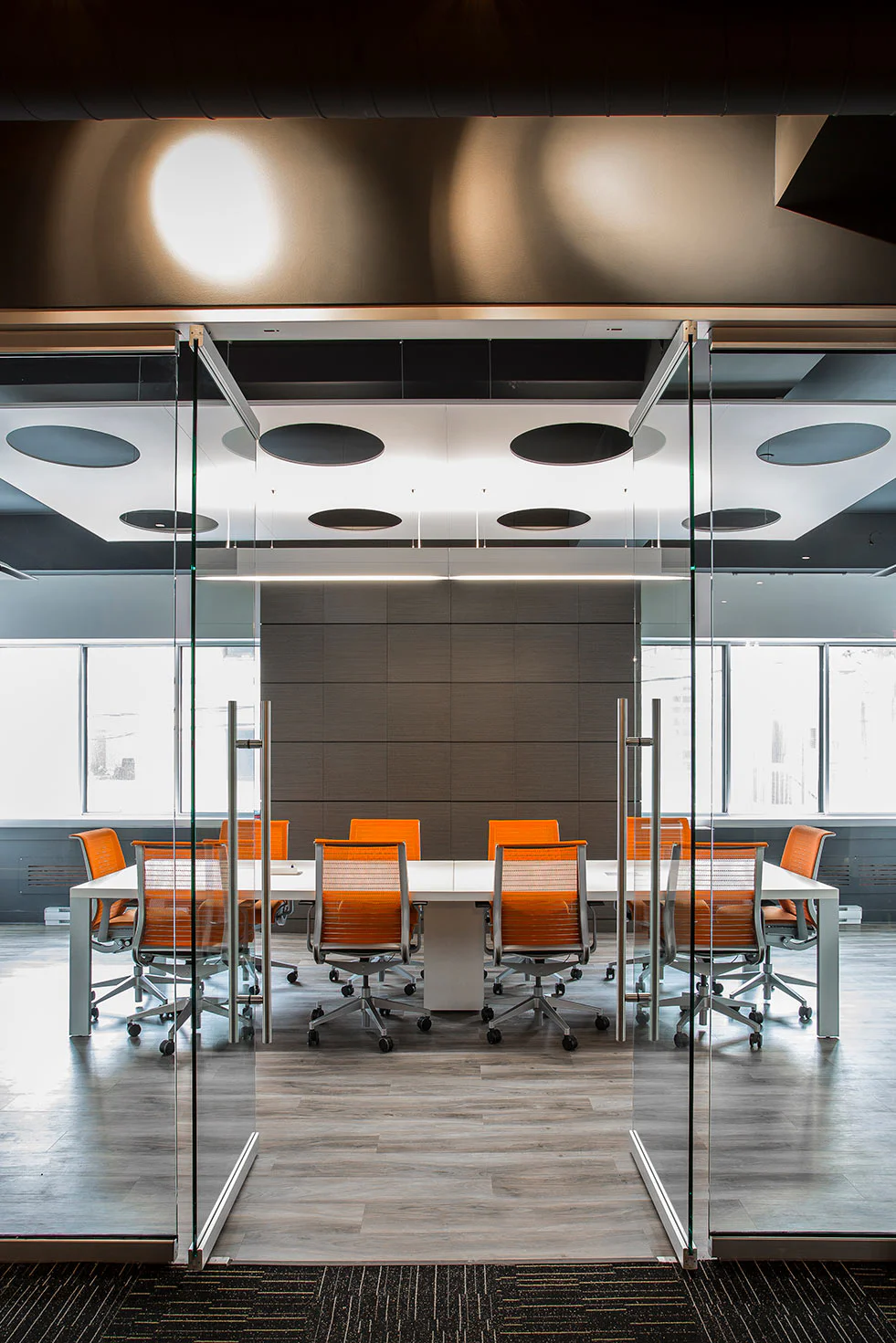 A modern conference room with a long white table, several orange office chairs, glass doors, large windows, wood flooring, and a ceiling with round black acoustic panels.