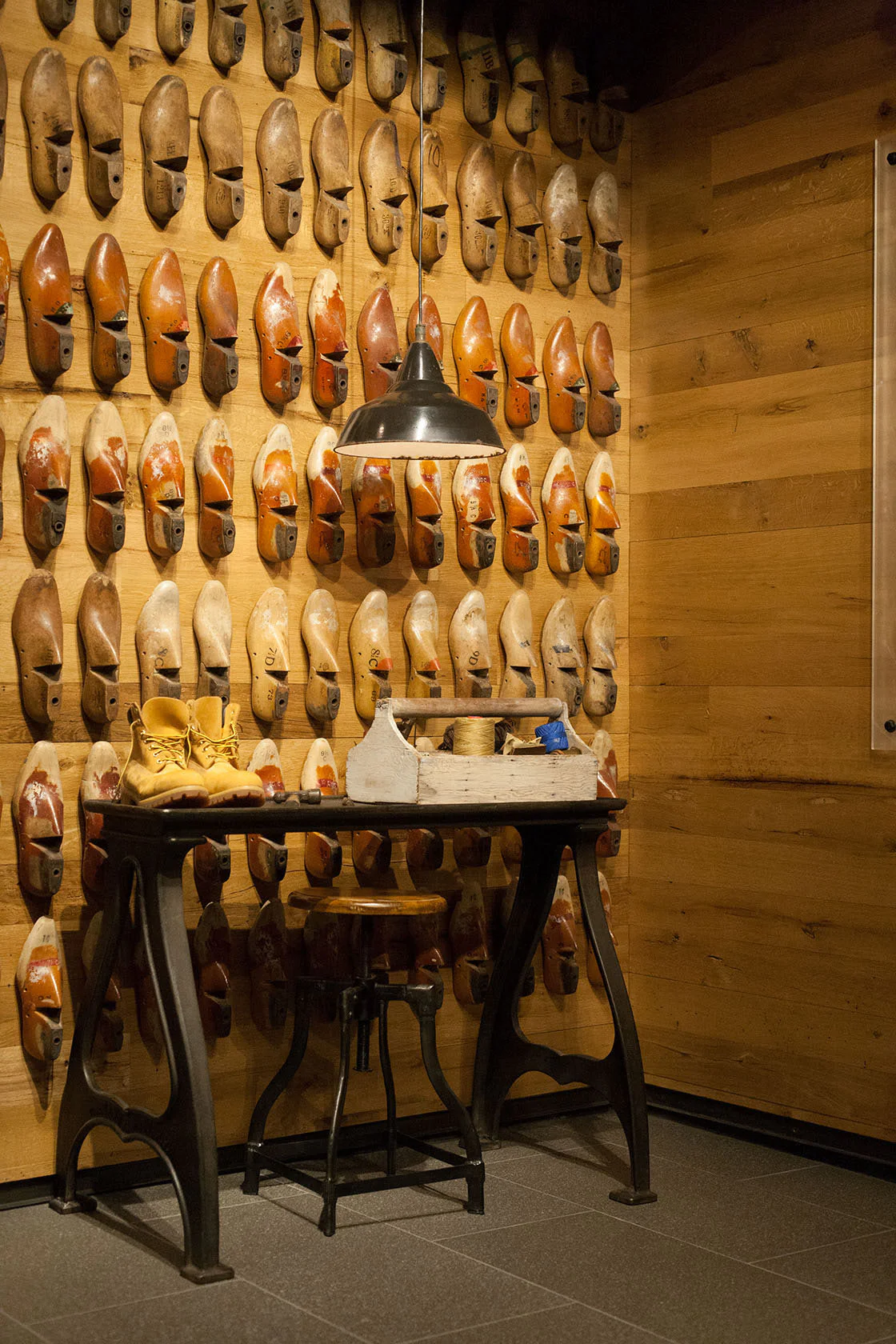 A wooden wall displays many vintage shoe lasts, with a black metal table below holding tools, yellow shoes, and a lamp hanging above. The setting suggests an old-fashioned shoemaker’s workshop.