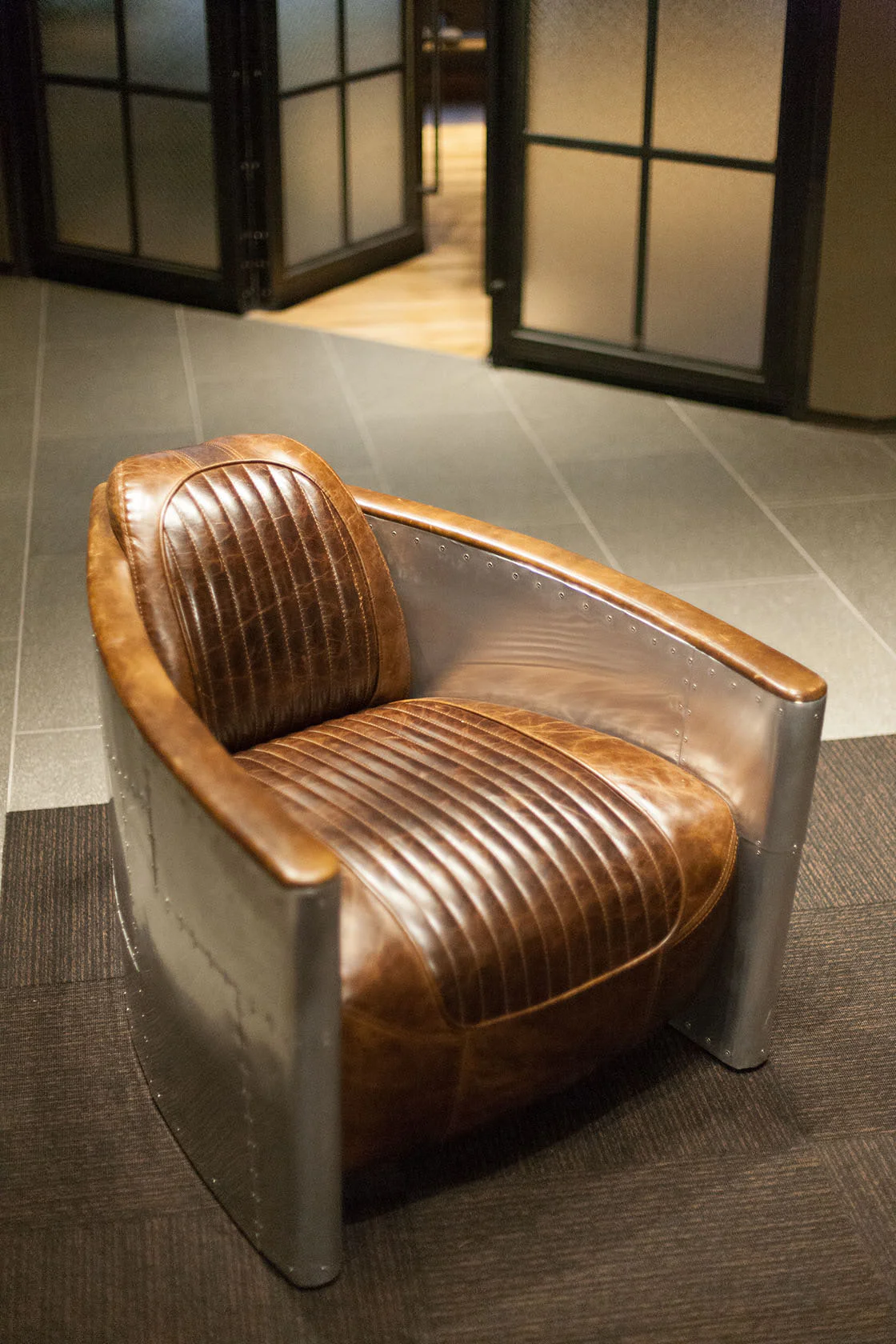 A modern armchair with brown leather cushions and ribbed stitching, set in a metal frame with rivets, sits on a carpeted floor in a room with glass and metal partition walls.