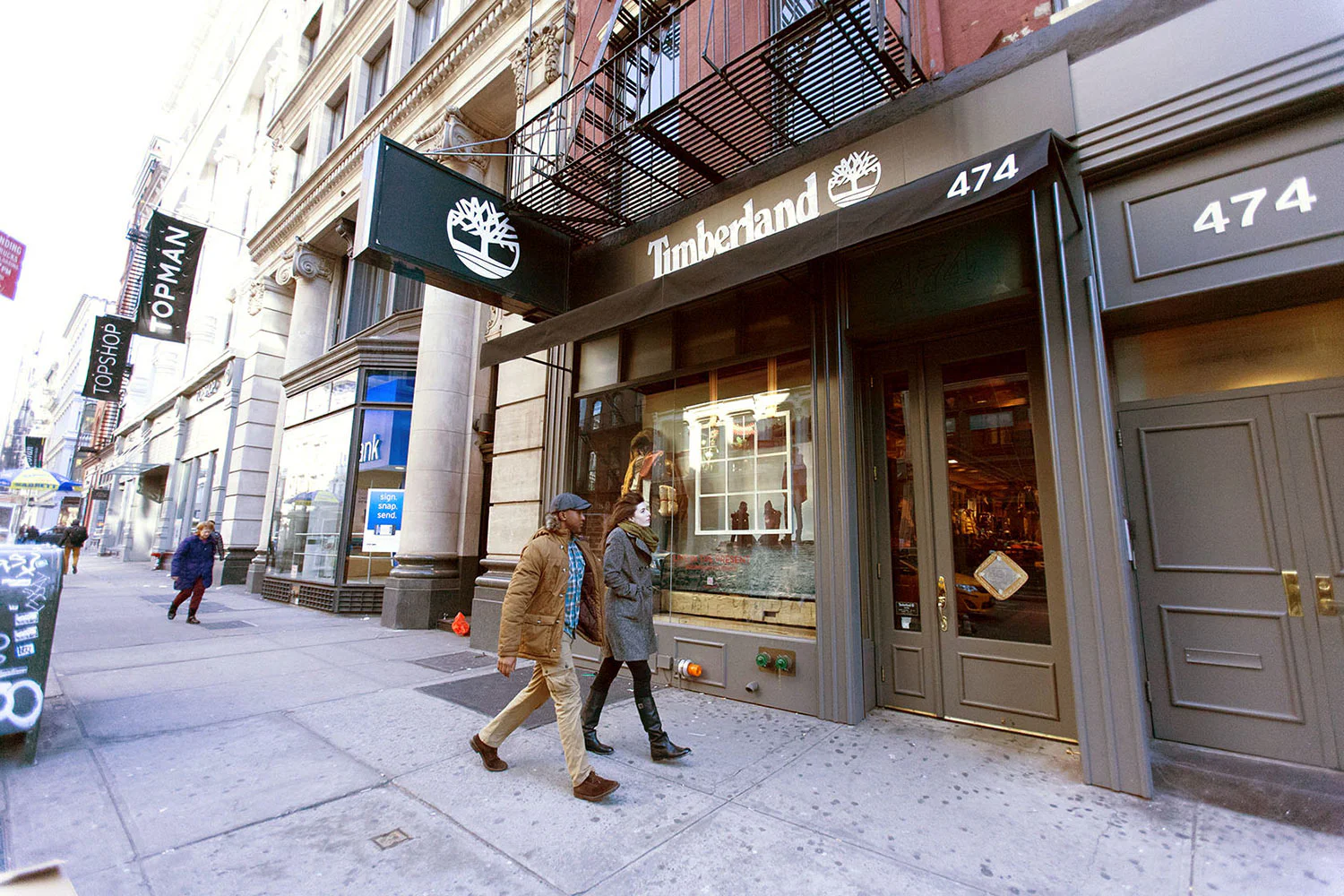 Two people walk past the Timberland store at 474 on a city street lined with shops and buildings on a clear day. The storefront displays the Timberland logo above the entrance.