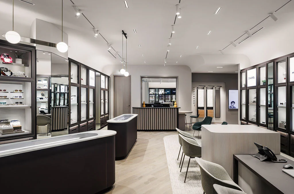 A modern eyewear store with sleek display cases showcasing glasses, a service counter, comfortable seating areas, light wood floors, and bright, even lighting throughout the stylish, spacious interior.