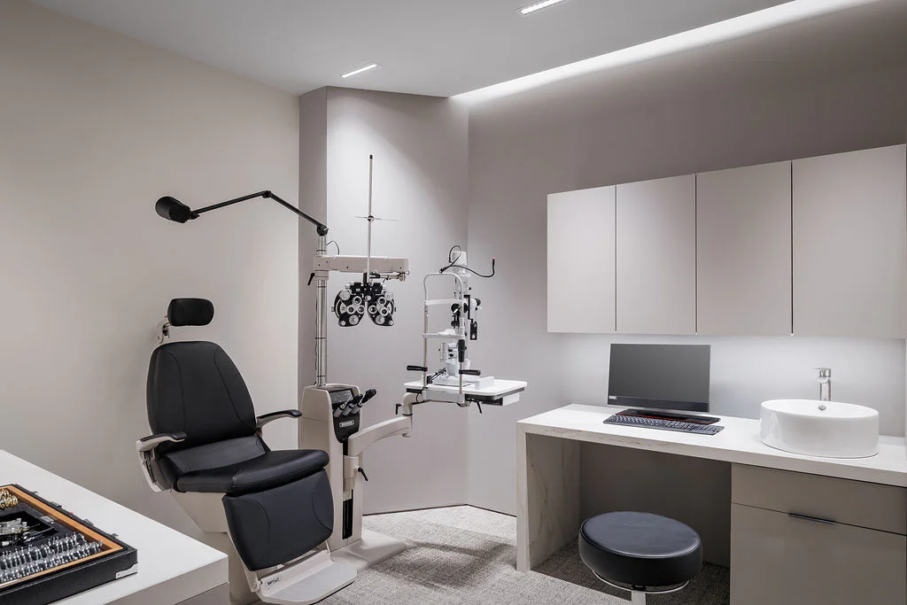 A modern optometry exam room with an eye exam chair, phoropter, counter with a sink, computer, and cabinets. The space is clean, minimal, and well-lit, with neutral colors and sleek design.