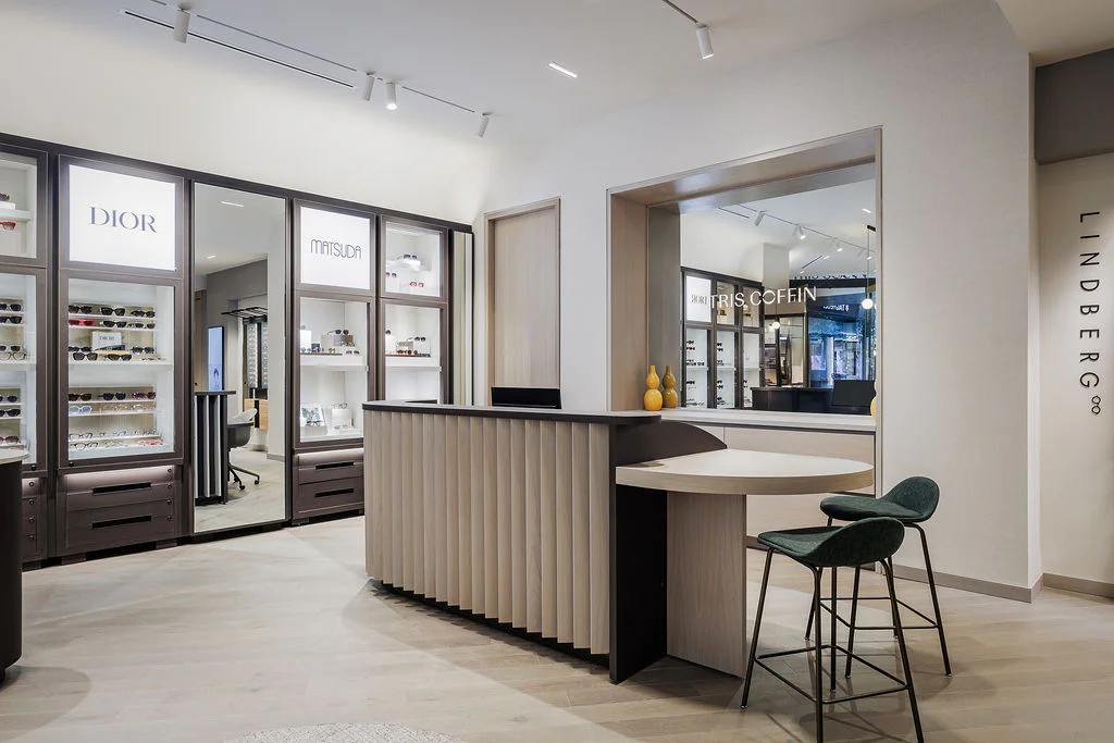 Modern optical store interior with sleek display shelves featuring designer eyewear brands like Dior, Matsuda, and Lindberg. A curved reception desk and two green chairs are in the foreground under soft lighting.