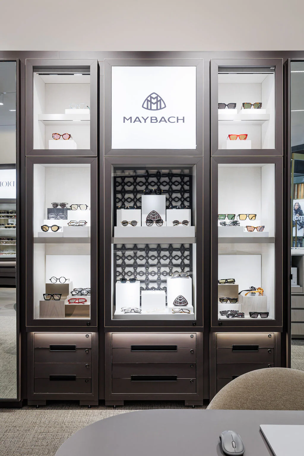 A display case featuring various luxury eyeglasses and sunglasses by Maybach, arranged on multiple shelves with bright lighting and a prominent Maybach logo at the top center.