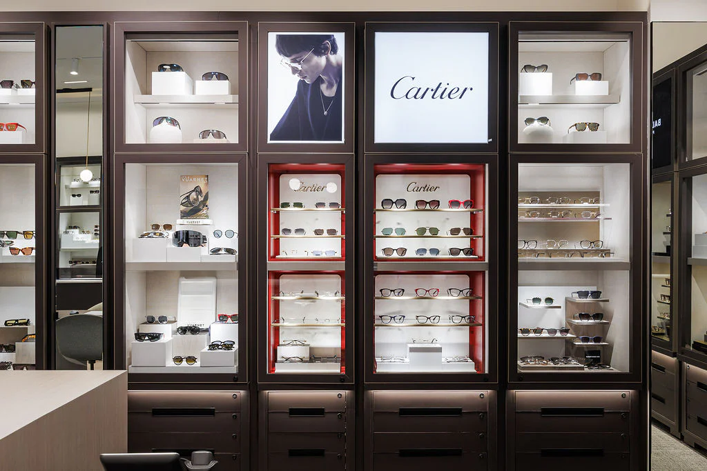 A luxury eyewear store display features various Cartier eyeglasses and sunglasses arranged on illuminated shelves, with branded signage and a stylish promotional image above the display.
