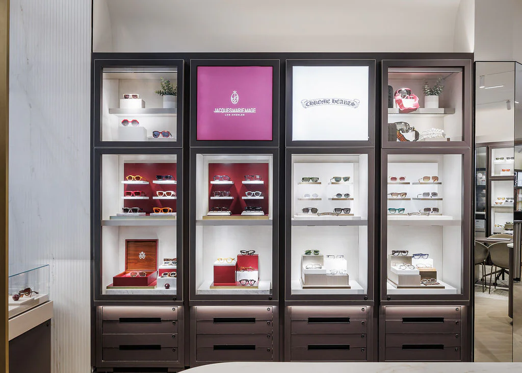 A well-lit display with shelves showcasing various designer sunglasses in a modern eyewear store. The cases feature different styles and colors, with brand logos highlighted on illuminated panels above the shelves.